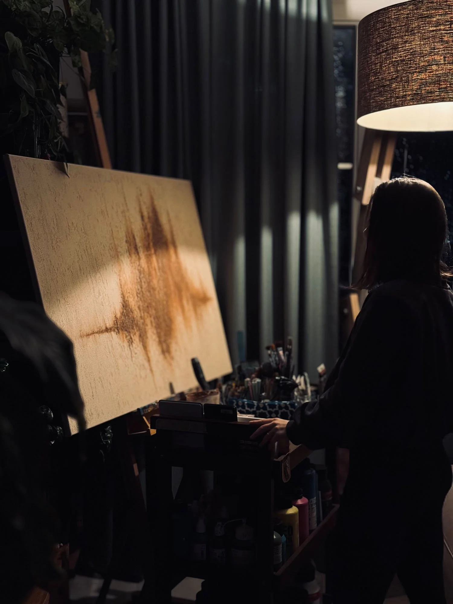 Person standing in front of an easel with a painting in an art studio, illuminated by a floor lamp, with art supplies and dark curtains in the background.