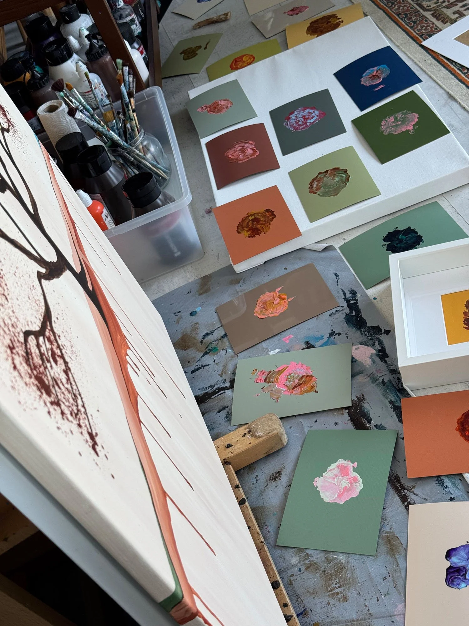 Assorted small paintings of flowers on colored paper laid out on the floor and a work table with paint bottles, brushes, and a canvas.