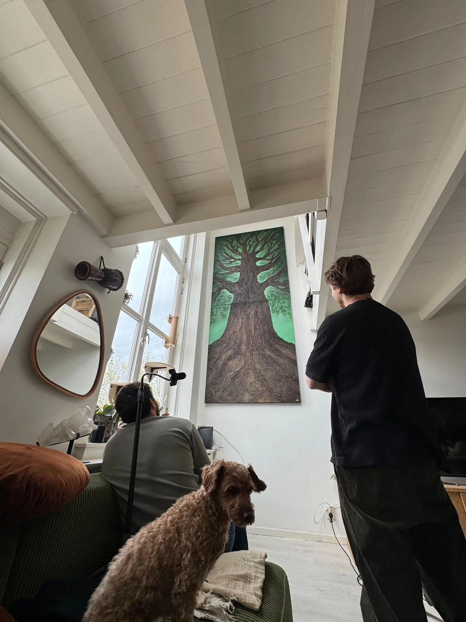 Interior of a bright living space with a large framed tree painting on the wall, ceiling beams, a mirror, and multiple people including a woman sitting on a green couch with a large dog in the foreground and a man standing nearby, engaged in conversation.