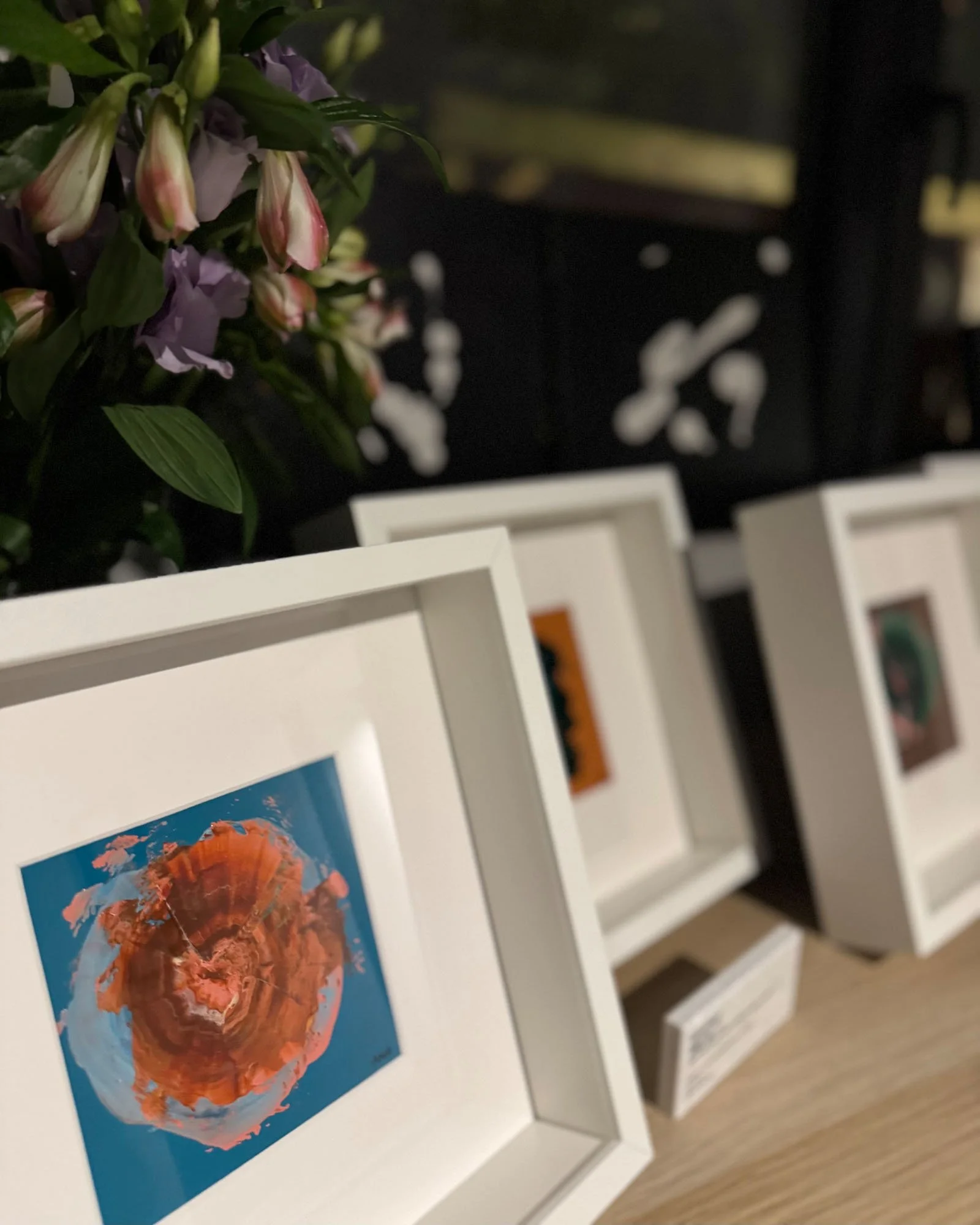 A white display shelf with framed artwork, one of which features an abstract orange and red flower on a blue background. The background includes a dark wall with shelves holding various items.