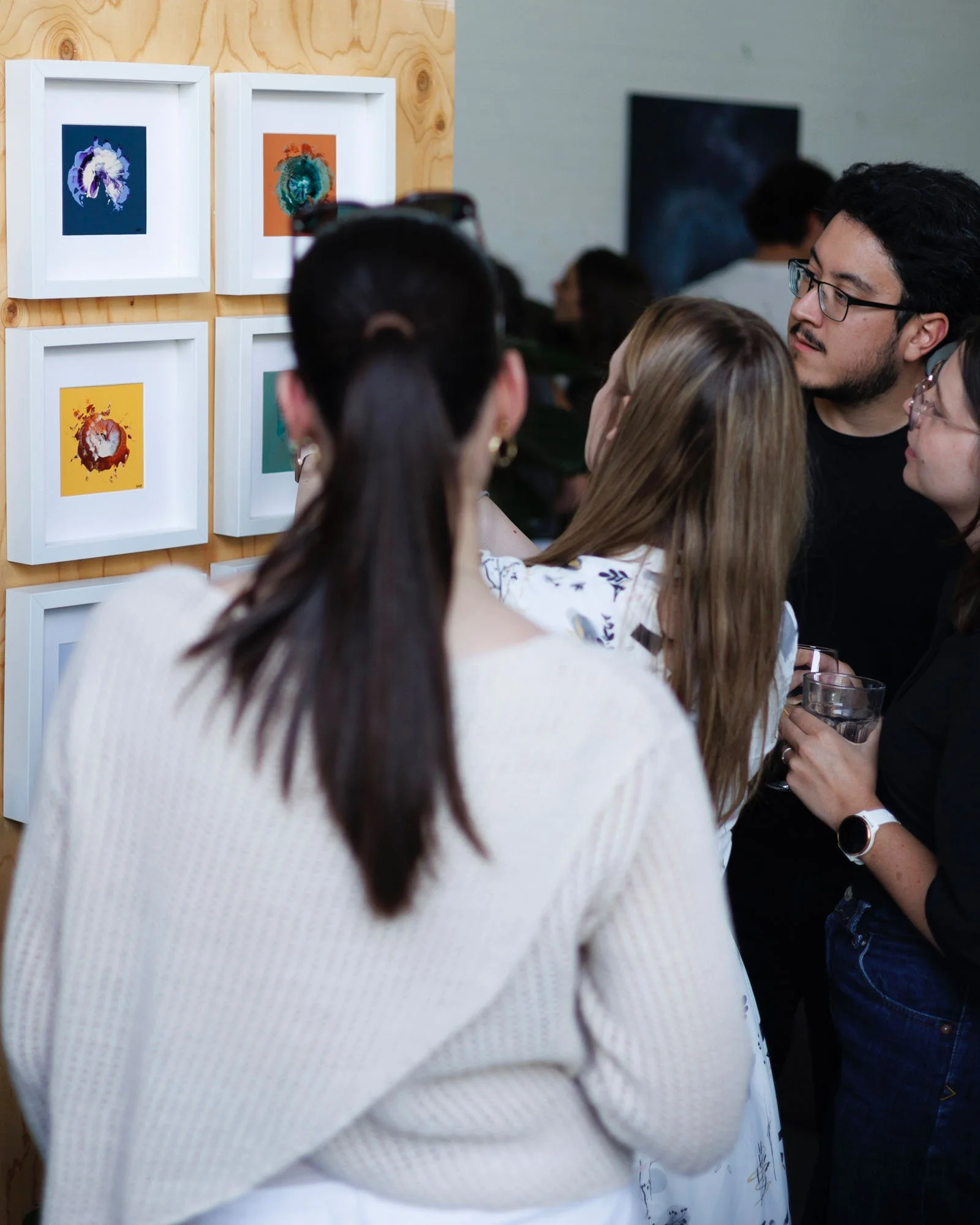 People gathered at an art exhibition viewing colorful abstract paintings displayed on a wall with a wooden background.
