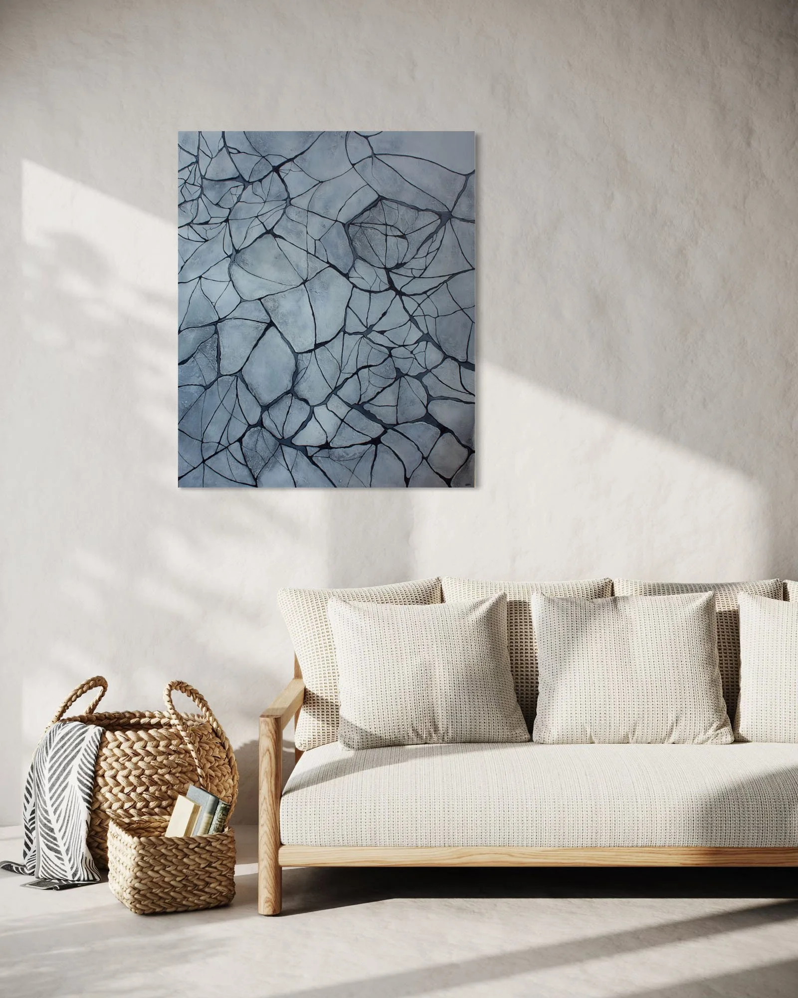A minimalist living room with a white textured wall, a modern painting of cracked ice or marble, a cream sofa with textured pillows, a woven basket with books, and a wooden armrest.