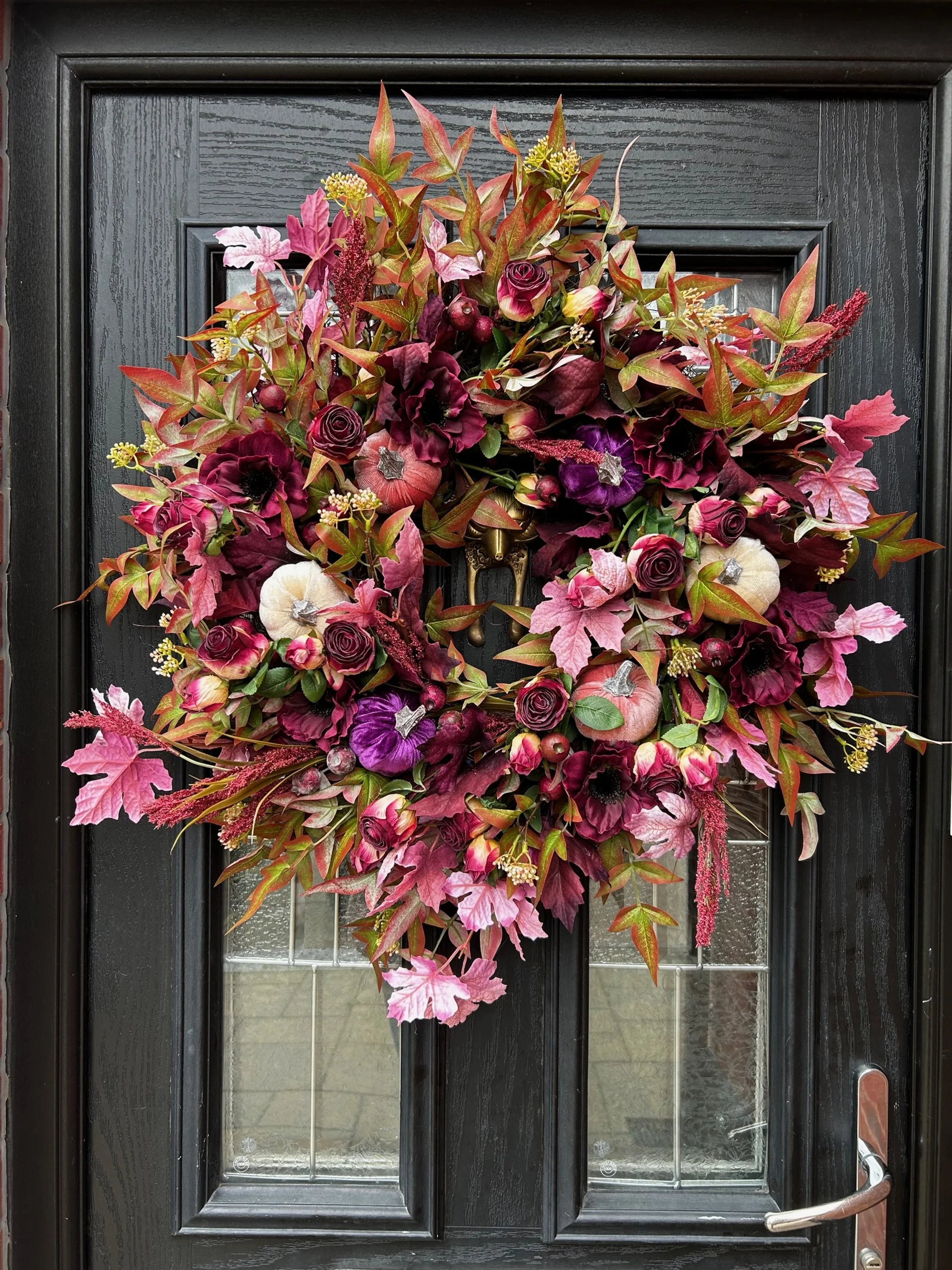 A decorative fall wreath on a black front door, featuring pink, purple, and white flowers, red and green foliage, small pumpkins, and silver accents.