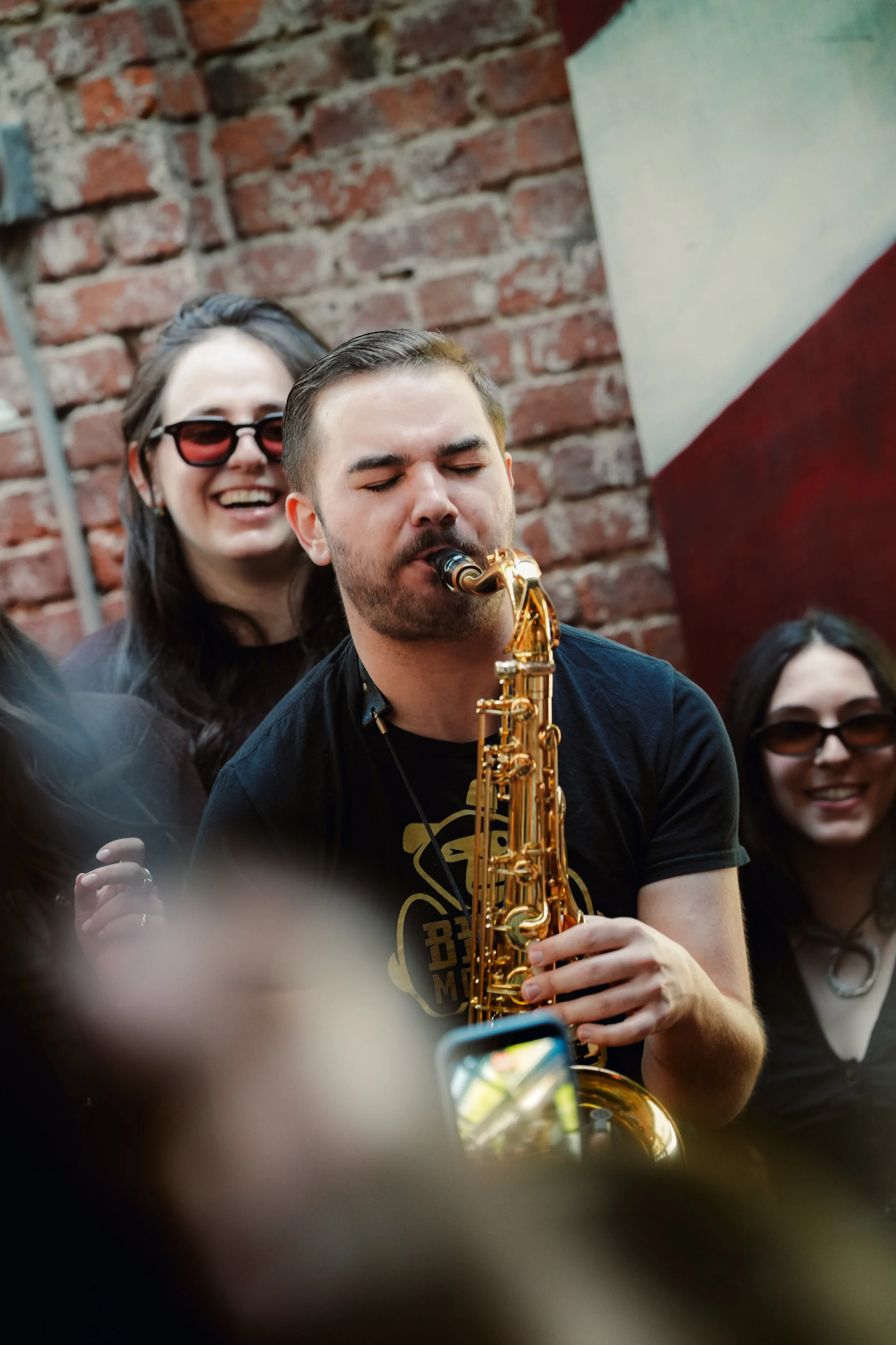 A group of musicians performing together in a cozy, brick-walled venue with large windows, including a singer holding a microphone, a guitarist, a saxophonist, and a woman with sunglasses.