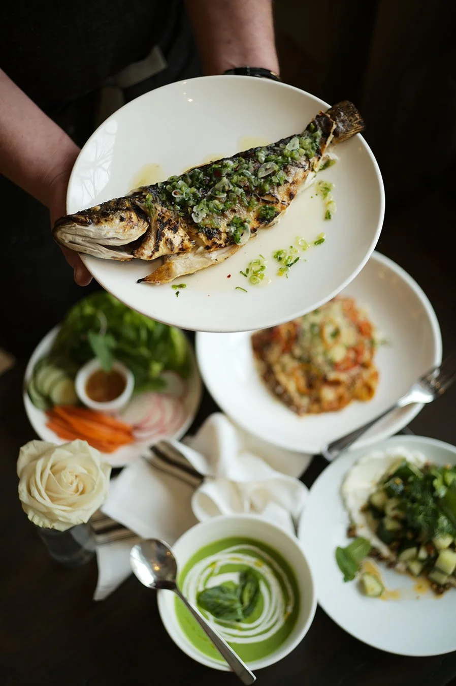 Restaurant table filled with food with someone holding a plate with a grilled marinated trout with a sautéed  garlic and herb sauce.