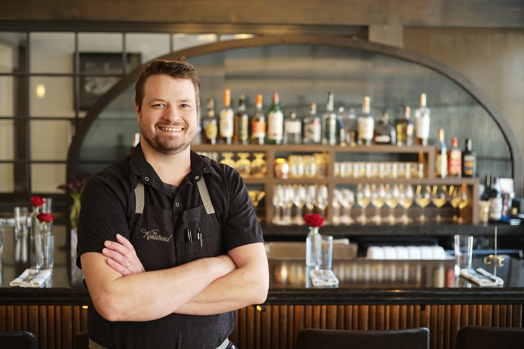 Chef Joel standing in The Kenwood dining room in front of the beautiful bar with curved glass backdrop.