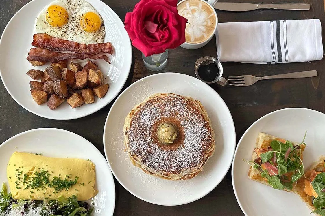 Full brunch spread with omelette stack of pancakes bacon eggs breakfast potatoes focaccia benedict and a latte.