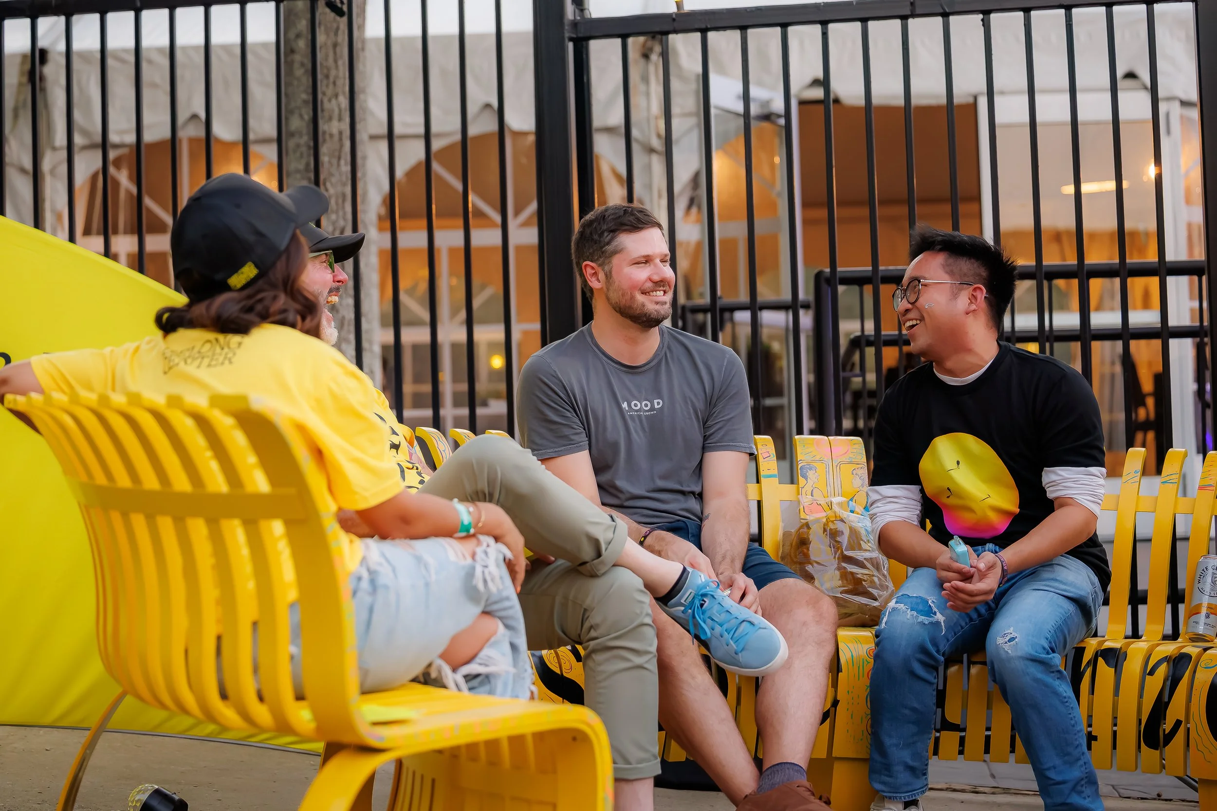 A group of four young people sitting and talking on yellow chairs outdoors, behind a black metal fence. They are smiling and appear to be enjoying a casual conversation in a lively setting.