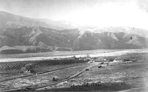 Historical black and white photograph of a rural landscape with mountains in the background, open fields, and a few buildings or farms.
