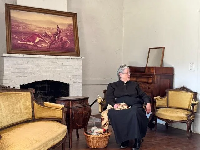 An elderly woman sitting on a vintage-style armchair with a yellow upholstered seat and back, wearing a black dress. She is holding a small item on her lap and looking to her right. To her right is another matching vintage armchair, and in front of h