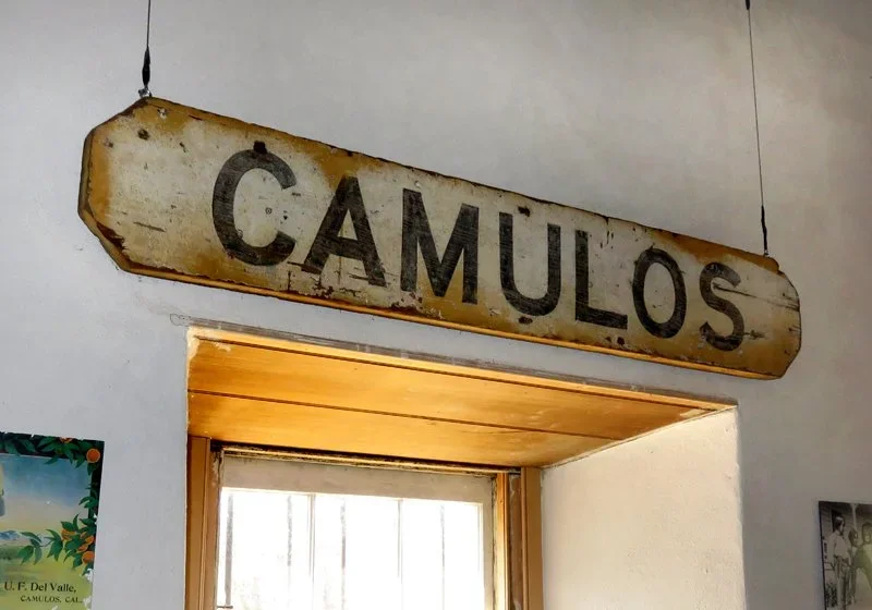 Rusty vintage sign with the word "CAMULOS" hanging above a doorway with a wooden ceiling, in a building with white walls.