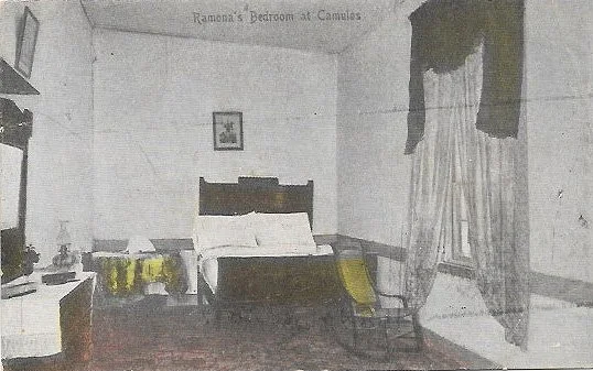 Vintage black and white photo of Ramona's bedroom at Camulos, featuring a bed, a small table with lamps, a chair, and curtains by a window.