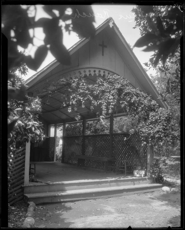 Black and white photo of a small outdoor church pavilion with a cross on the gable, surrounded by trees and foliage.
