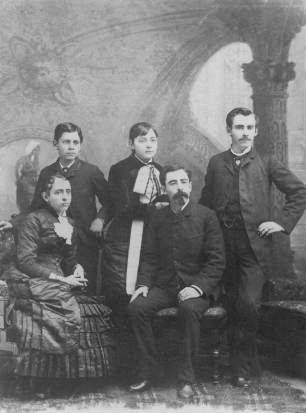 A black and white photograph of a family portrait with five people, including a seated man with a moustache, three standing children, and a seated woman in Victorian-style clothing inside a studio with painted background.