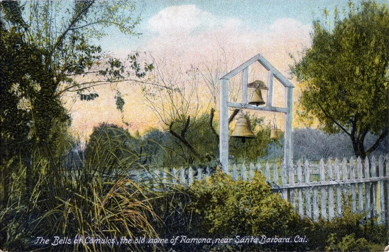 An outdoor scene at sunset featuring a white picket fence, trees, bushes, and wild grasses. A large outdoor bell with smaller bells hangs from a wooden frame. The caption reads: "The Bells of Camulos, the old home of Ramona, near Santa Barbara, Cal."