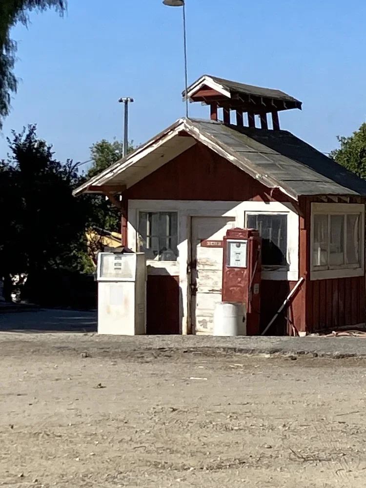 Small rustic building with roof, a red mailbox, and a white post box, surrounded by dirt ground and trees, under a clear blue sky.