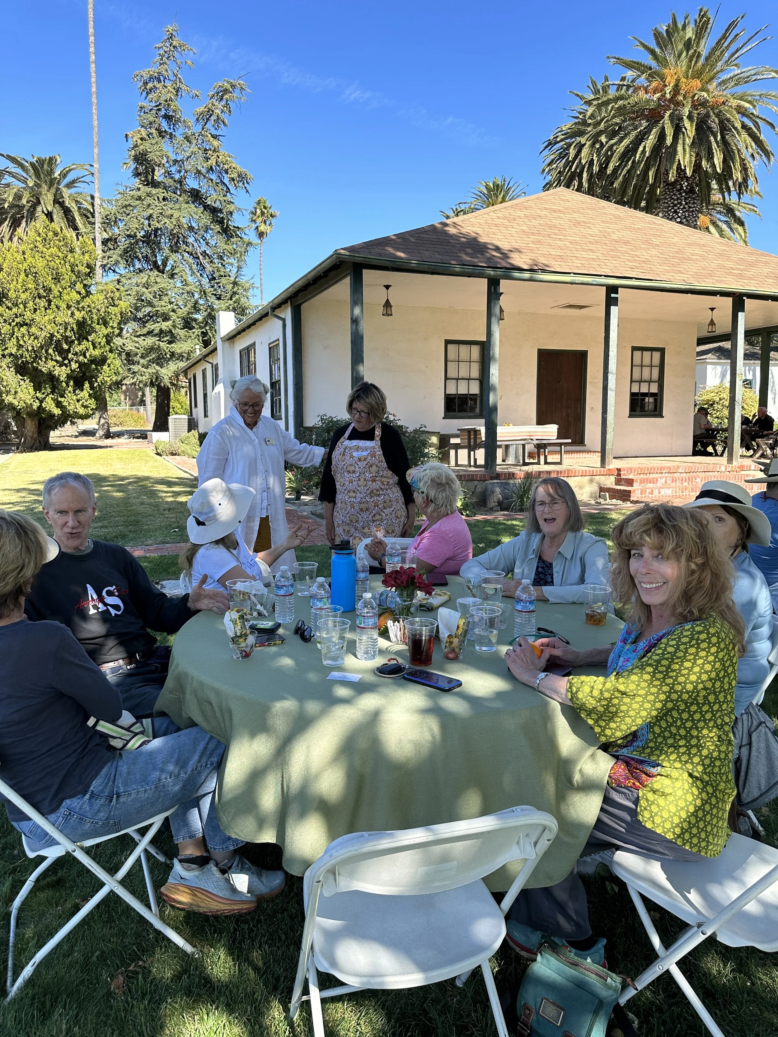 A group of people enjoying a outdoor gathering around a round table with a green tablecloth, set with water bottles and drinks, under a tree. Some are sitting and some are standing and talking in front of a historic building with palm trees in the ba