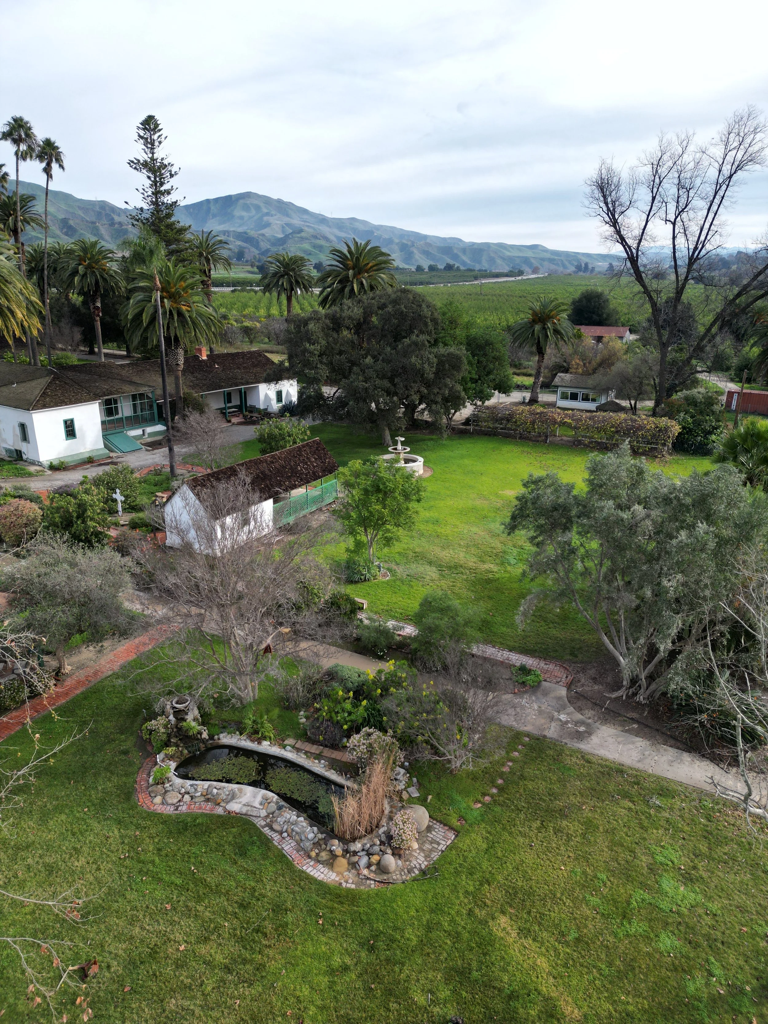 Aerial view of a lush green yard with a pond, trees, and a variety of houses and buildings, surrounded by mountains and a cloudy sky.