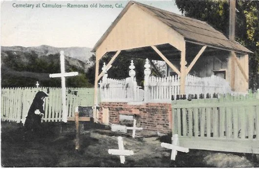 Historical cemetery scene at Ramona's old home in California, featuring a wooden shelter, white picket fence, crosses, and a person kneeling in prayer.