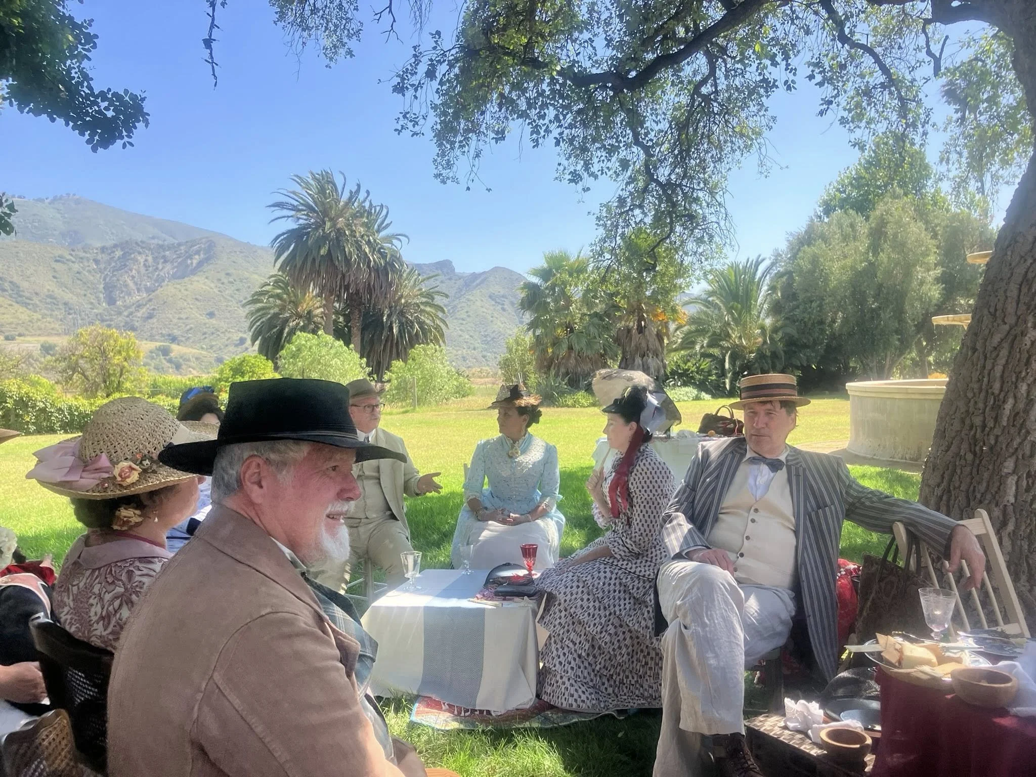People dressed in vintage clothing having a picnic outdoors under a large tree, with mountains and palm trees in the background.