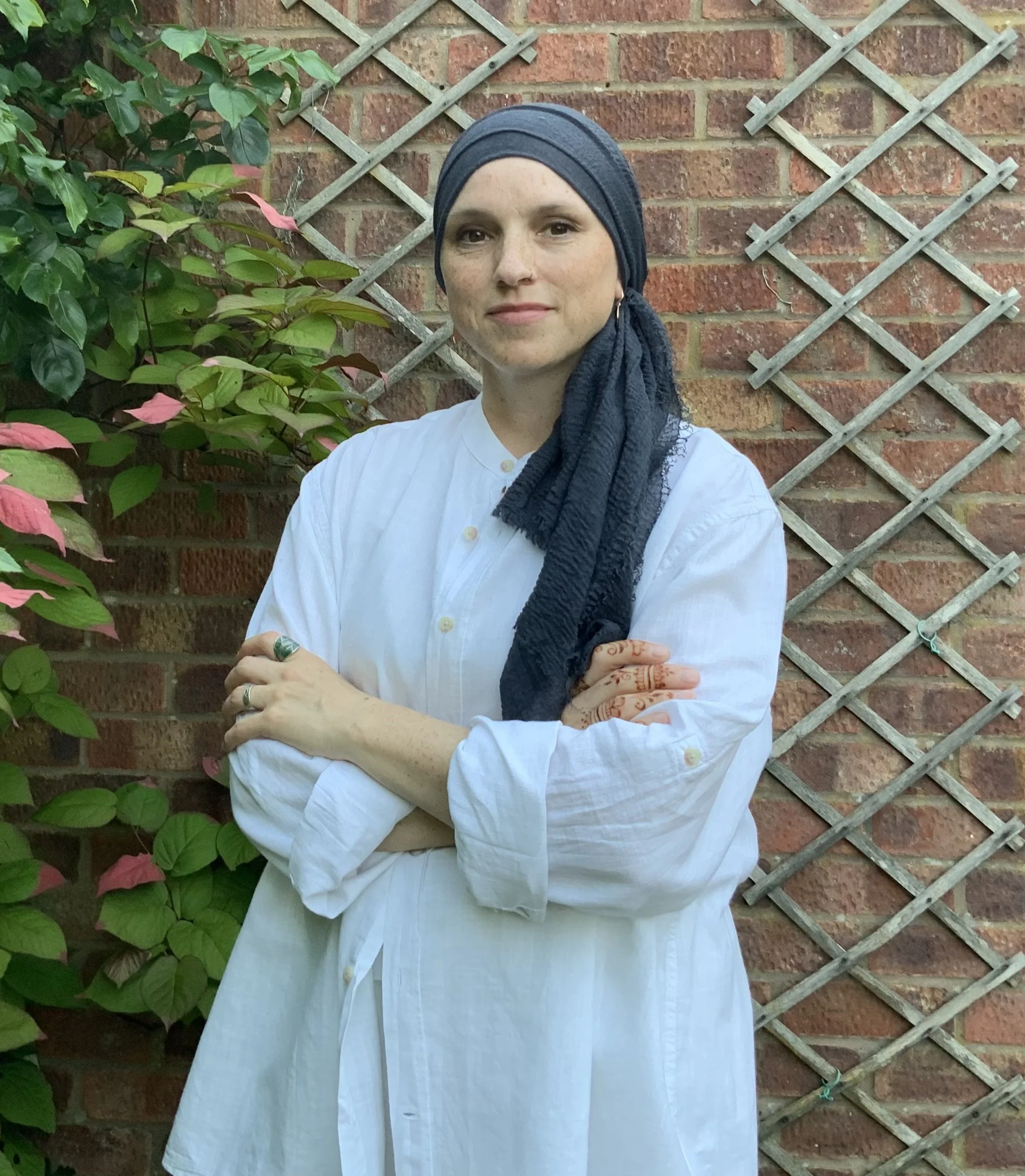 A woman in a white shirt and a dark headscarf standing outdoors against a brick wall with a wooden lattice, surrounded by greenery and pink flowers.