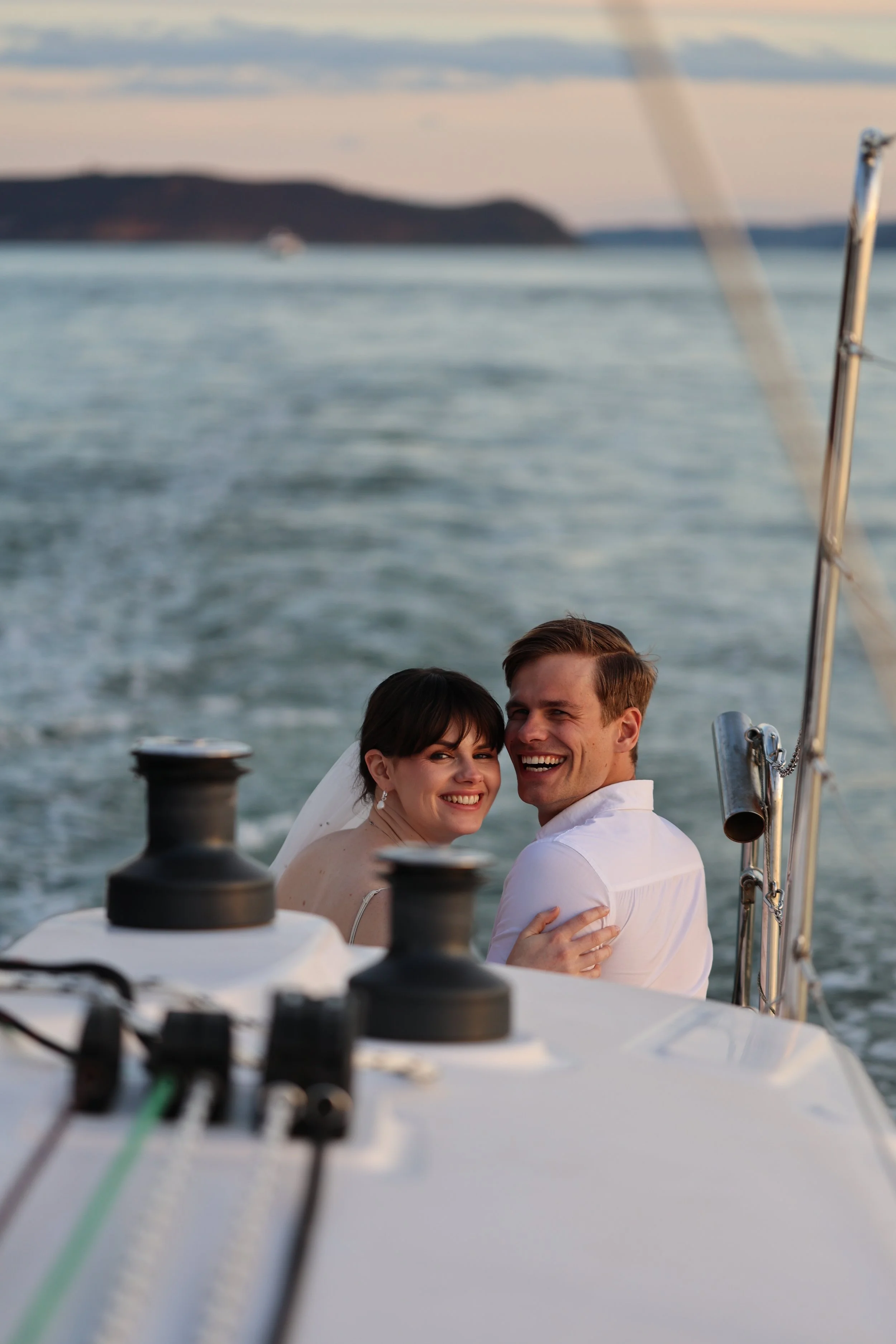 A happy bride and groom embrace on a boat with the water and distant shoreline in the background during sunset.