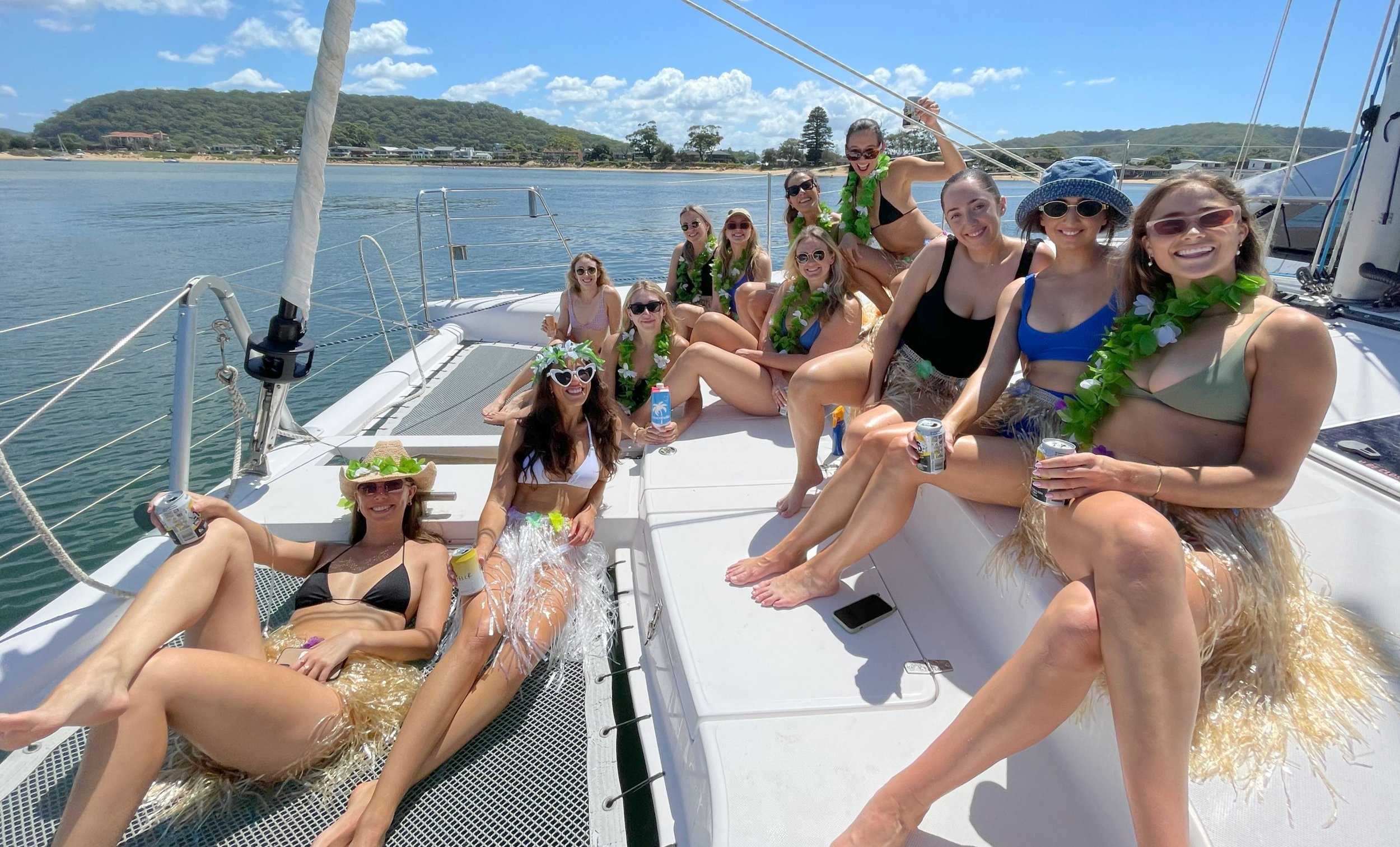 Group of women in swimwear and leis enjoying drinks on a sailboat with scenic coastline background.