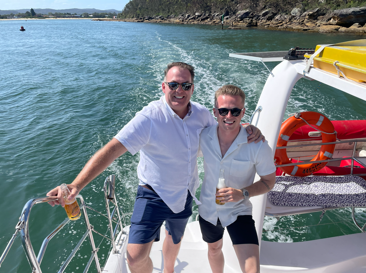 Two men with sunglasses smiling and holding drinks on a boat in a body of water, with rocky shoreline and trees in the background.