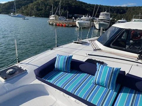 Part of a boat with a cushioned seating area with striped blue and green pillows, docked at a marina with other boats in the background.