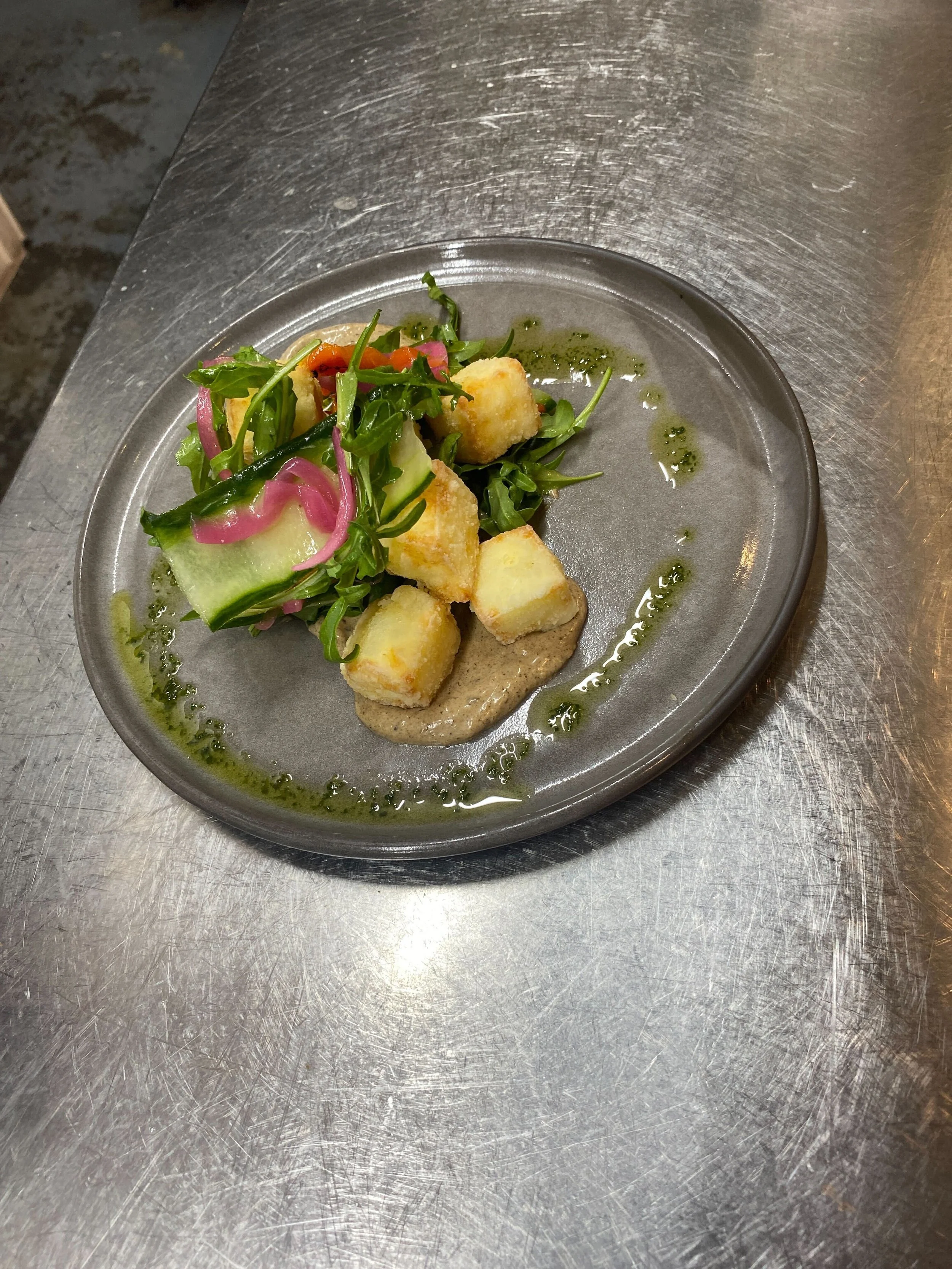A metal plate with a small salad of greens, cucumber, pickled onions, and croutons, topped with a green herb dressing.