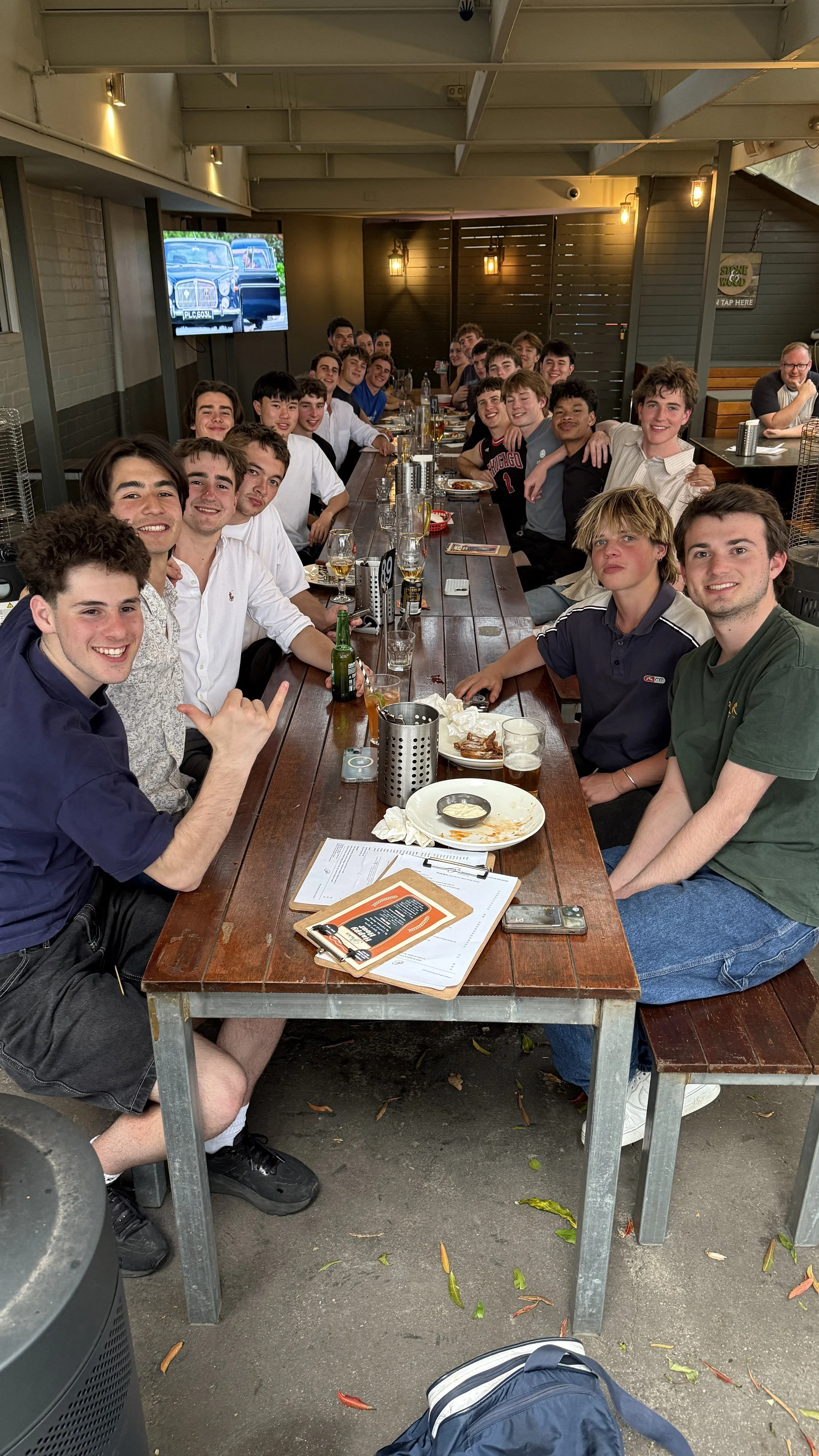 Group of young men sitting at a long wooden table in a restaurant or bar, smiling and enjoying food and drinks.