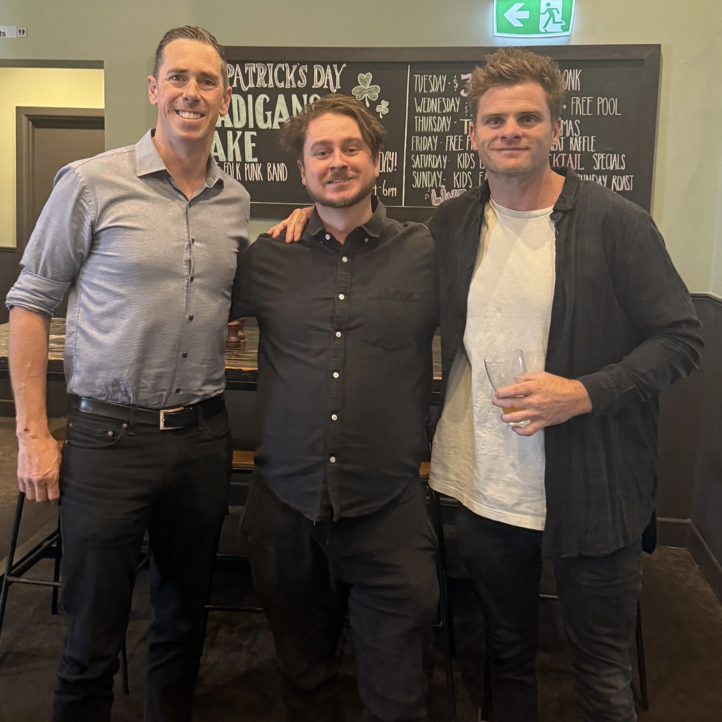 Three men standing together in a bar or restaurant, smiling at the camera. Behind them is a chalkboard sign with event details and a green emergency exit sign.