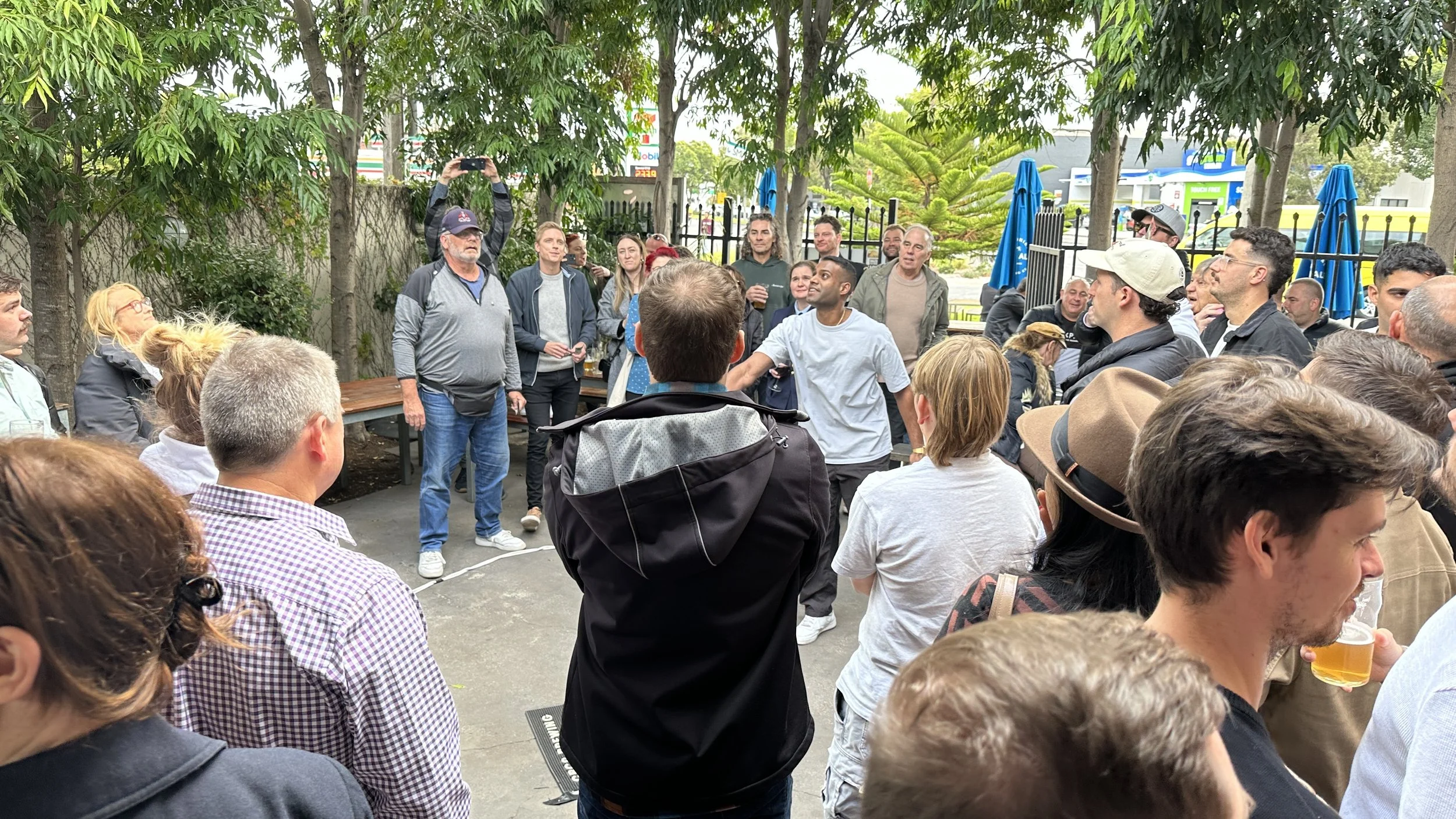 A group of people gathered outdoors, some standing and some sitting, playing 2-up on ANZAC day. The setting is the Palace Hotel beer garden with trees and outdoor tables.
