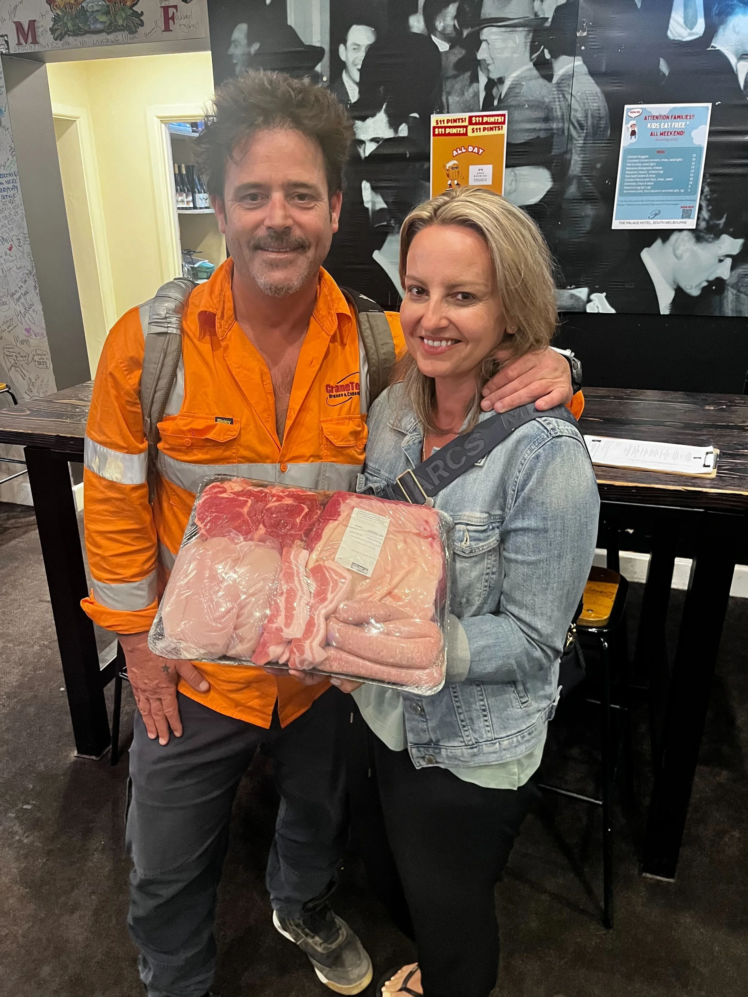 A man and woman standing together indoors, holding a meat tray they won. The man is wearing an orange work shirt, and the woman is wearing a denim jacket. They are smiling for the photo with a black and white mural in the background.