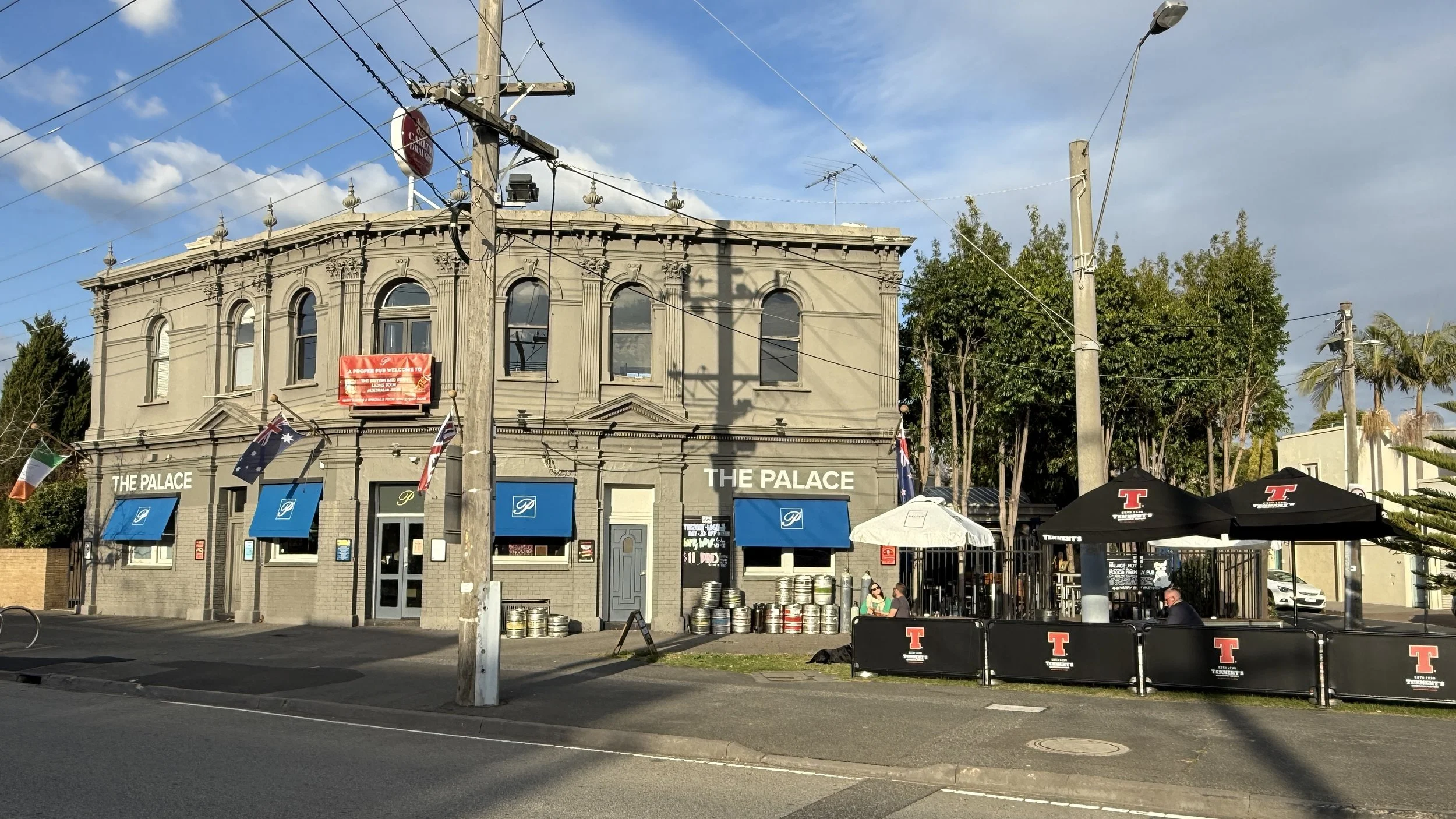 A two-story building named 'The Palace Hotel' with blue awnings and flags, situated on a street with outdoor seating under black umbrellas, some people sitting at tables.