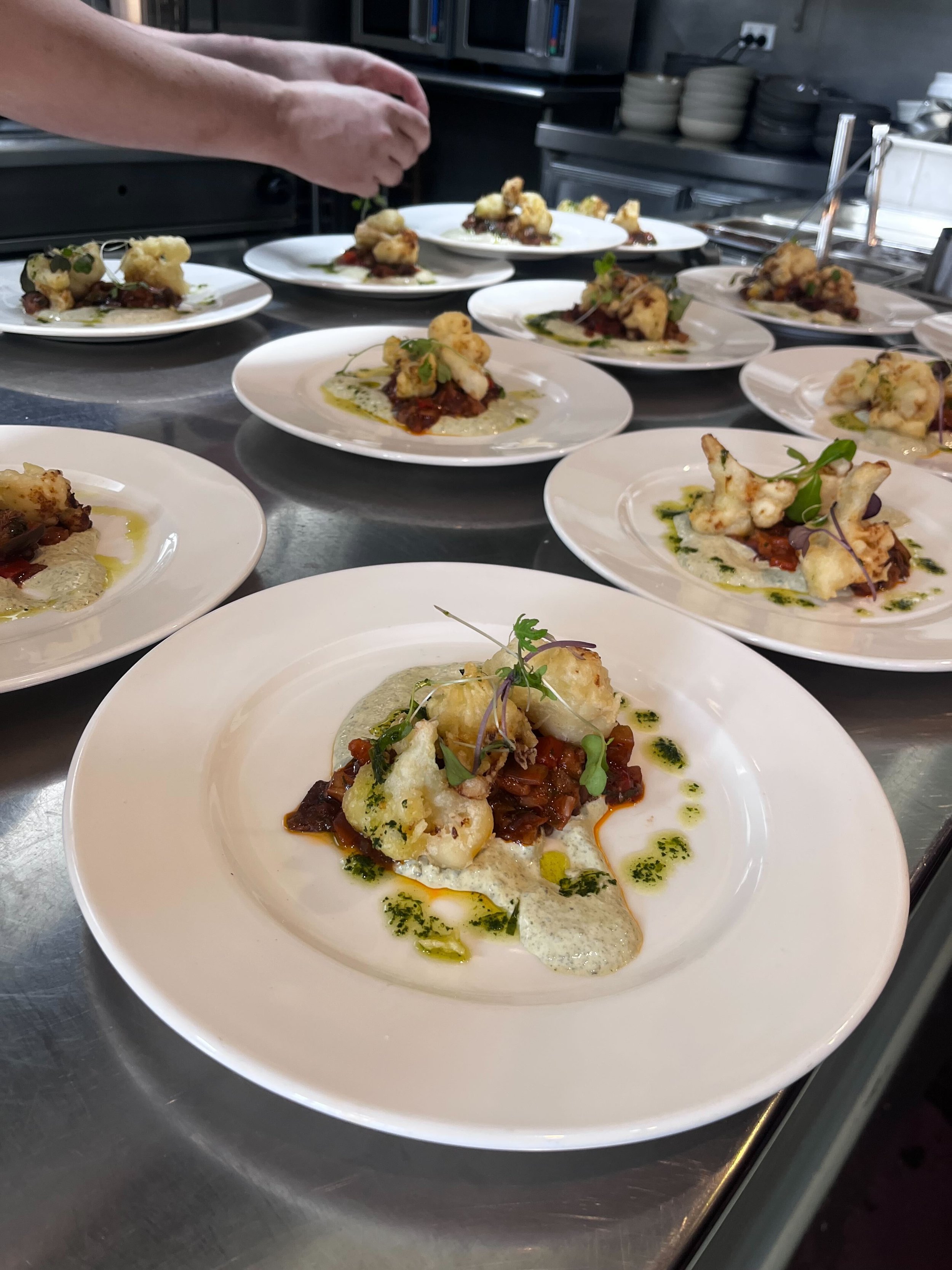 Plates of gourmet food with cauliflower, tomato-based sauce, microgreens, and sauce drizzled on top, set on a stainless steel counter in a professional kitchen.