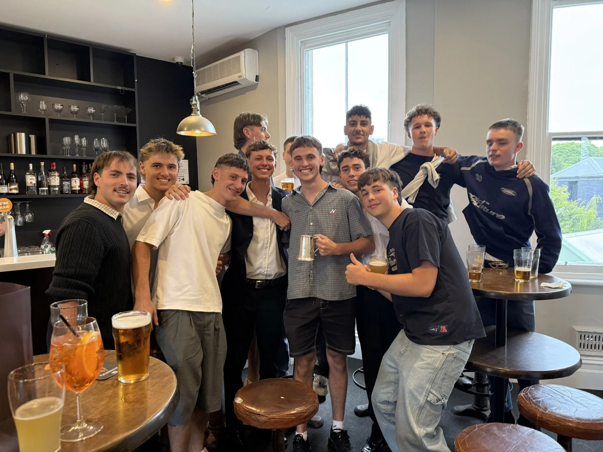 A group of young men upstairs at the Palace Hotel, smiling and posing for a photo, some holding drinks, with a black shelf of glasses and bottles behind them and a large window showing a green outdoor scene.
