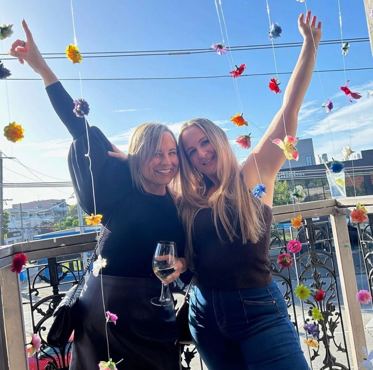 Two women smiling and celebrating outdoors on a balcony with colorful hanging flower decorations, holding a glass of white wine, under a clear blue sky.