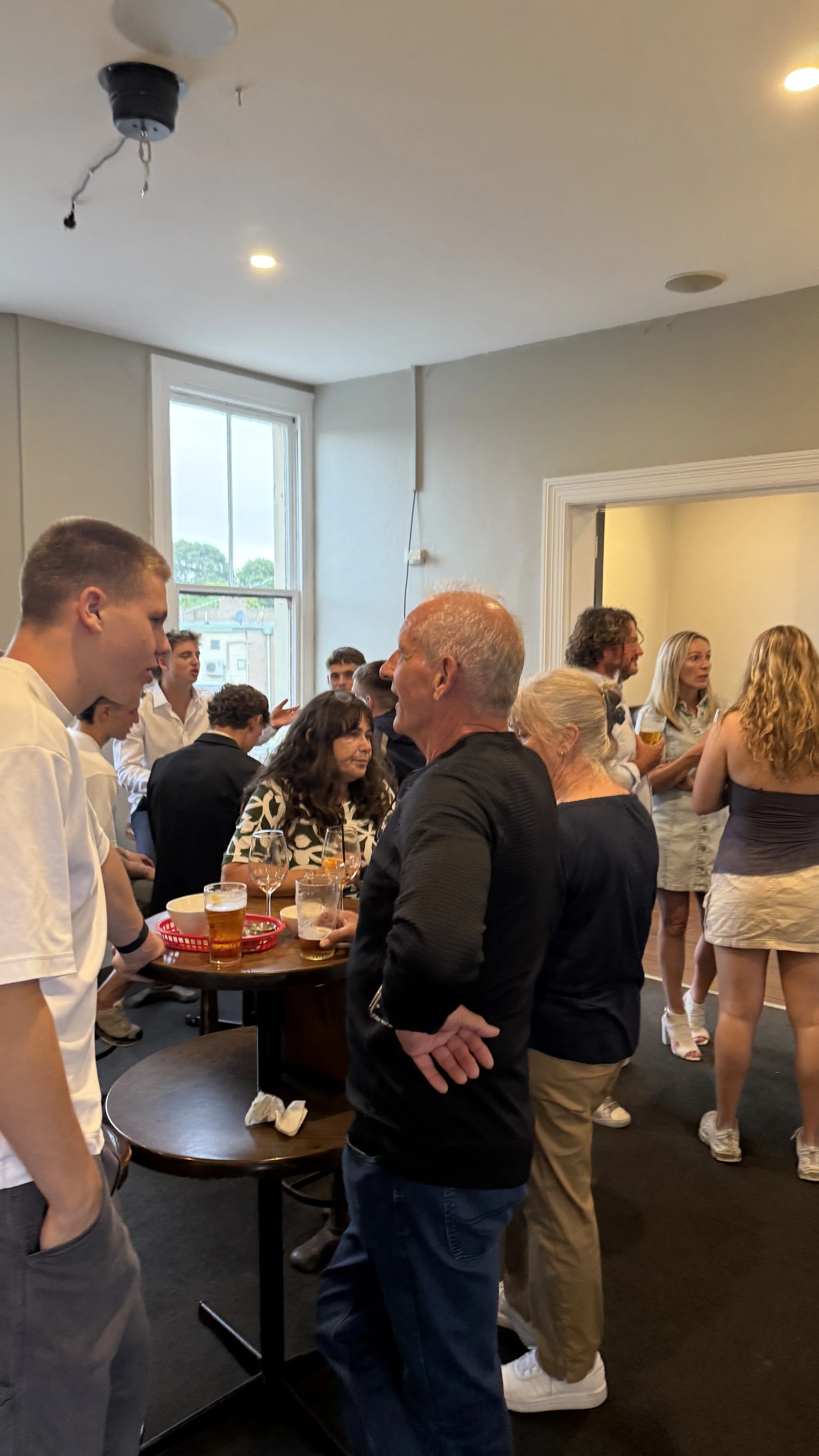 A group of people socializing upstairs at the Palace Hotel, standing around a small round table with drinks, with some standing near a doorway and others sitting or standing near a window.