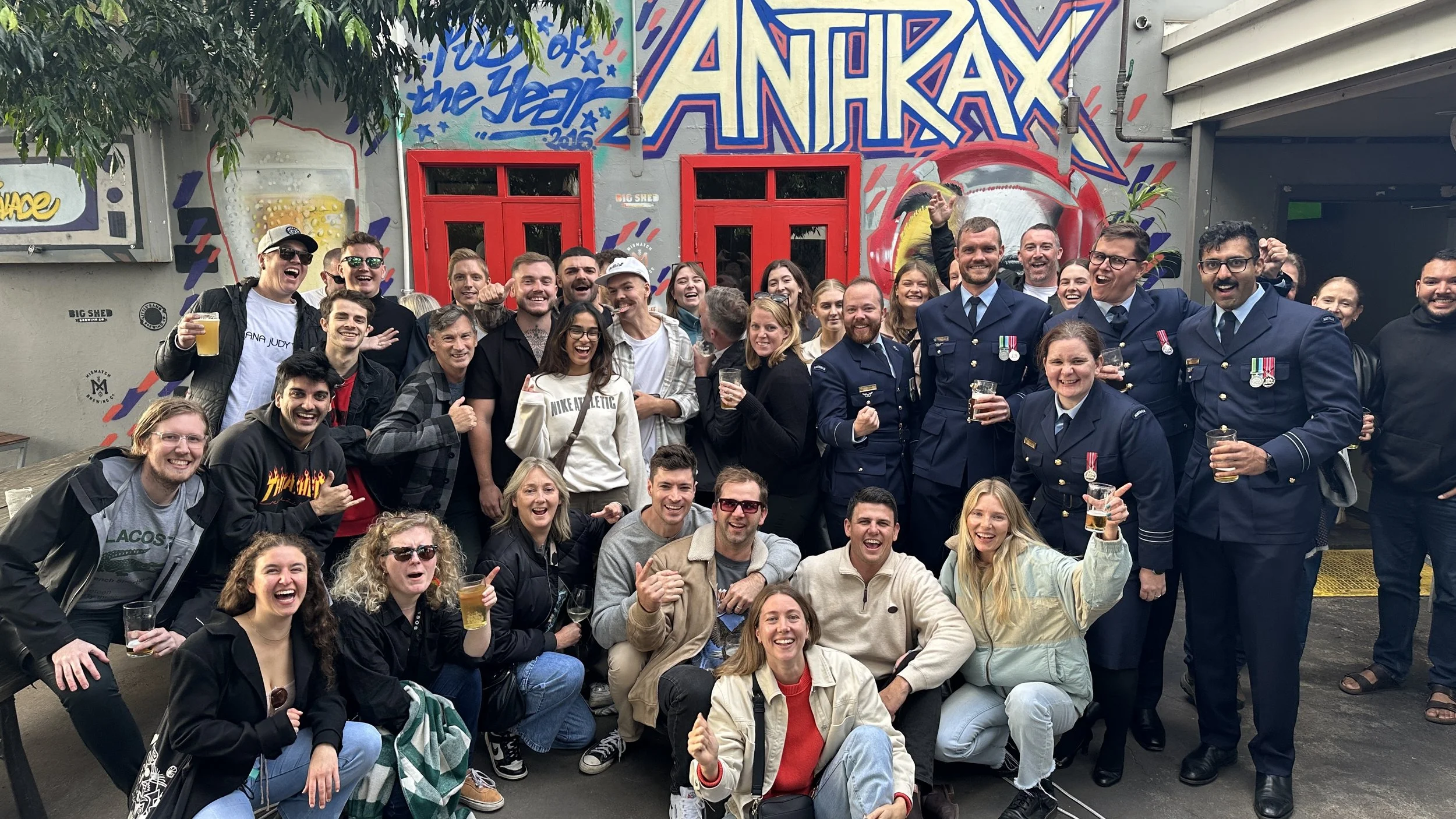 A large group of people, including civilians and military personnel, gathered outdoors for a celebratory photo. They are smiling, holding drinks, and some are making gestures. The background features a colorful mural with graffiti art and red windows