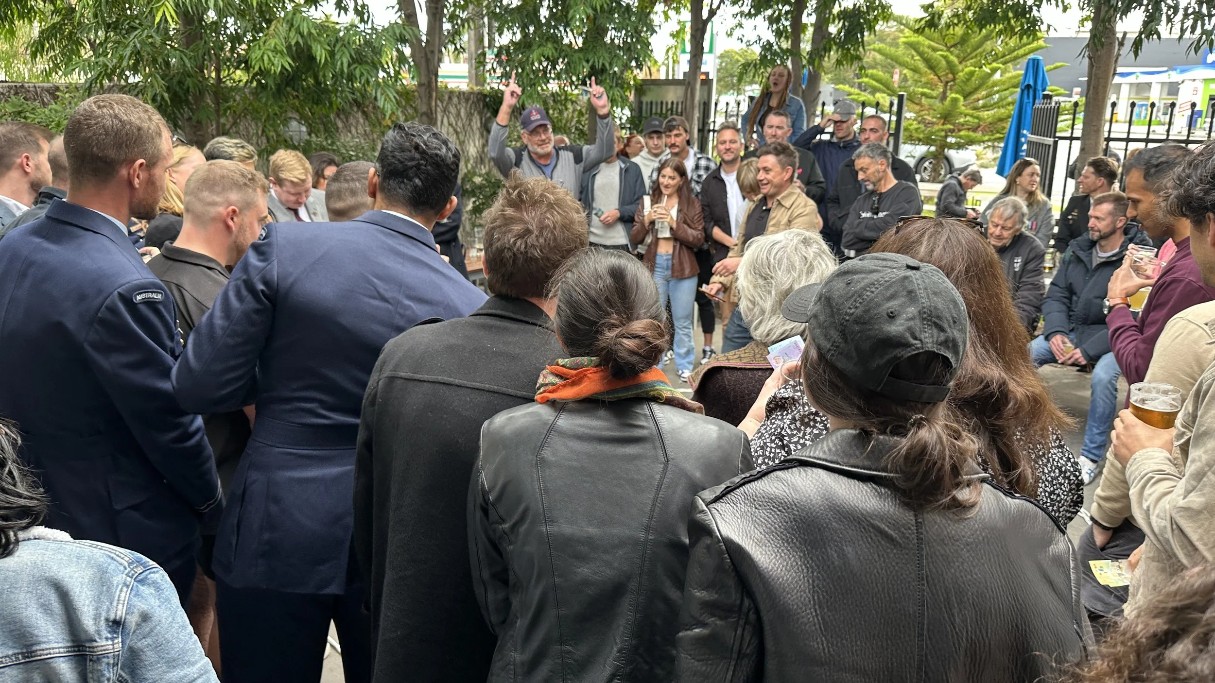 A group of people gathered outdoors playing 2-up on ANZAC day. The setting is the Palace Hotel beer garden with trees and outdoor tables and people watching and talking, holding drinks.