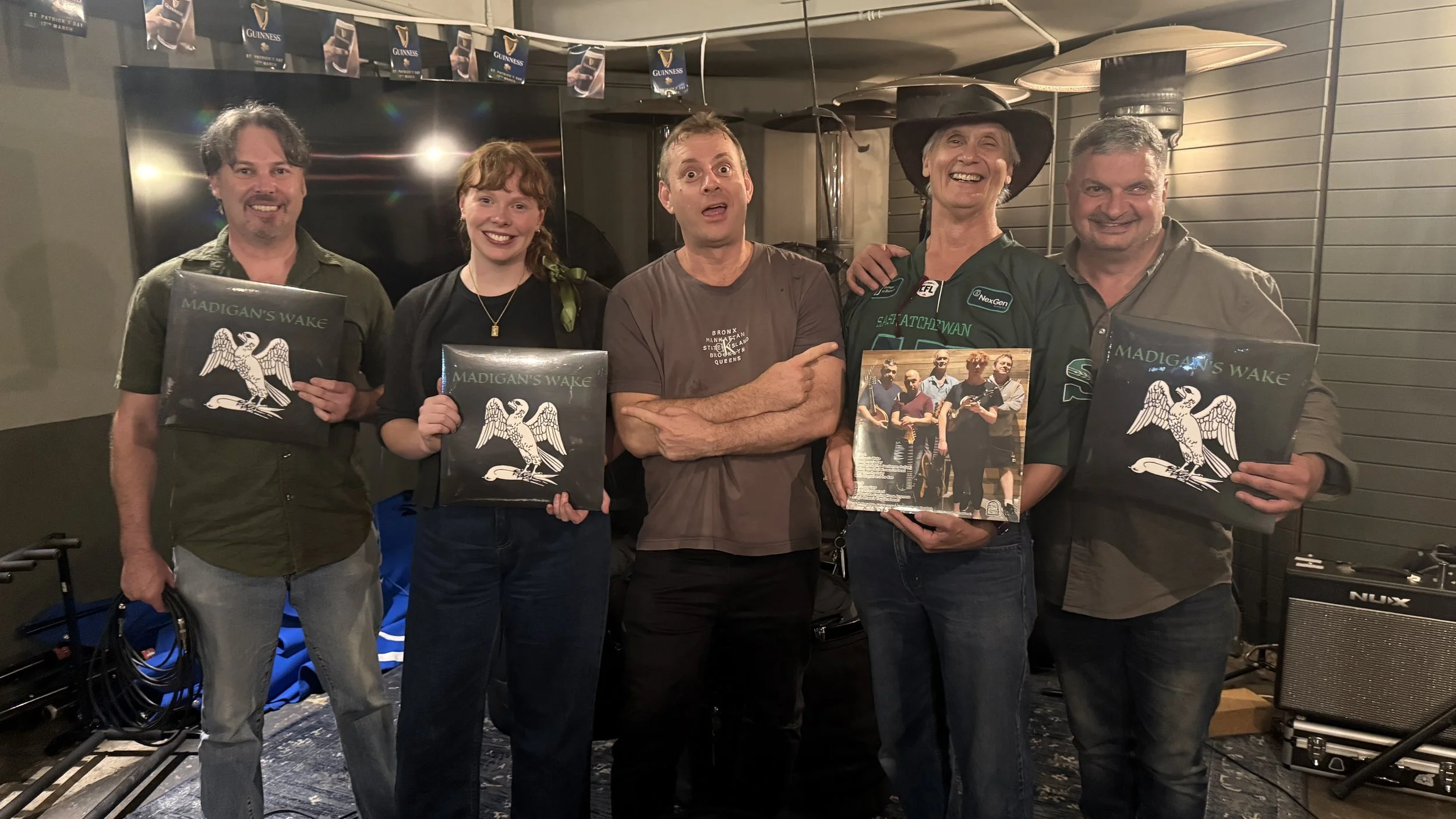 Five people standing together holding records and a picture, posing for a group photo, indoors with a Guiness banner and musical equipment in the background.