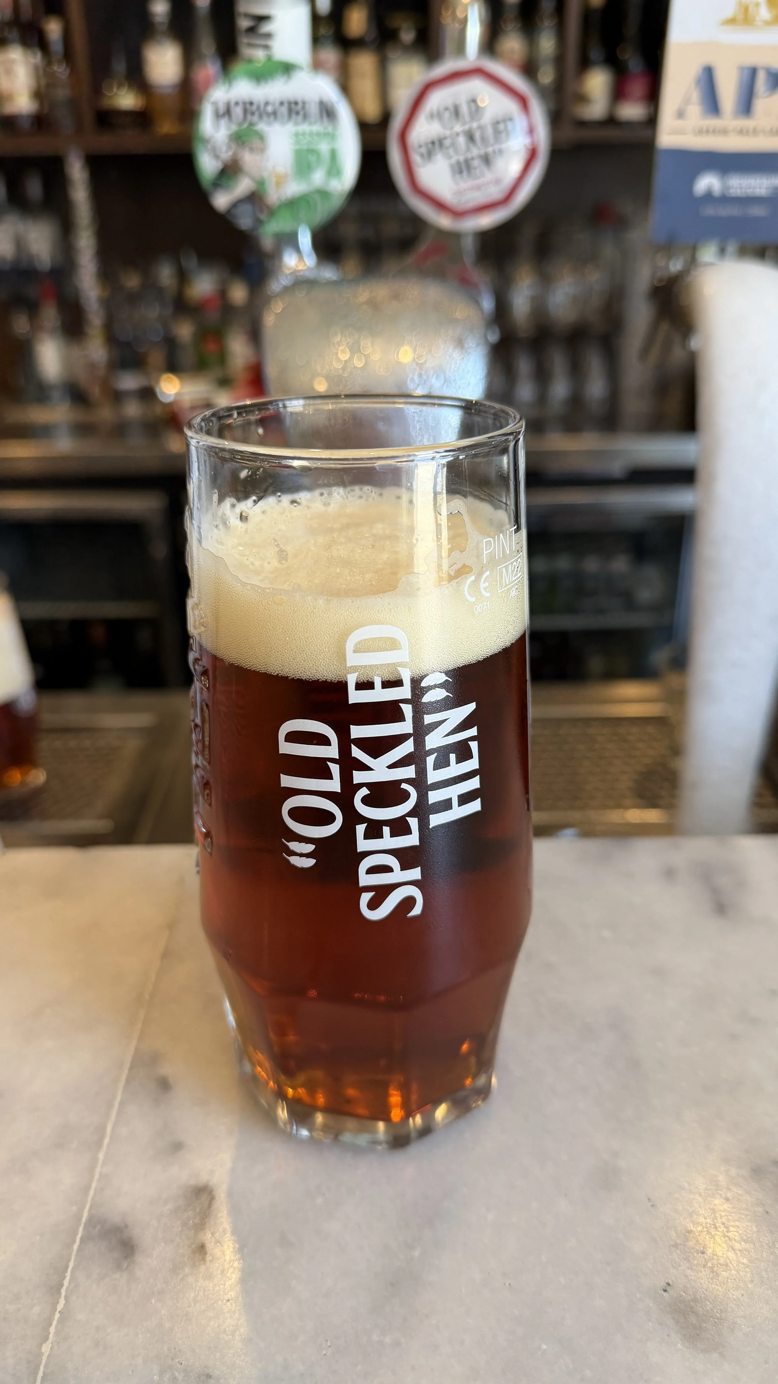 A pint glass of Old Speckled hen beer with a foamy head, featuring the quote 'Old Speckled Hen' in white text, placed on a bar or table with blurred bar taps and bottles in the background.