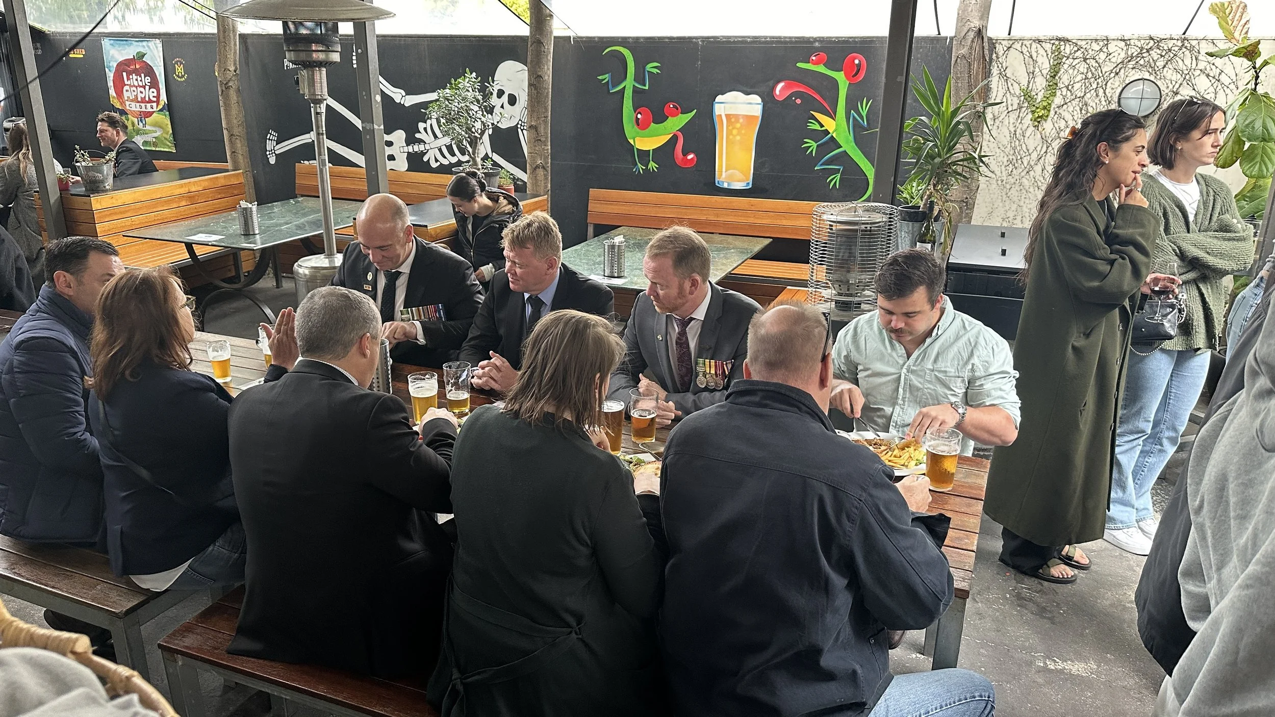 Group of people, including several men in military uniforms, sitting at a wooden picnic table, drinking beer and eating food, in beer garden with colorful murals, plants, and other patrons in the background.