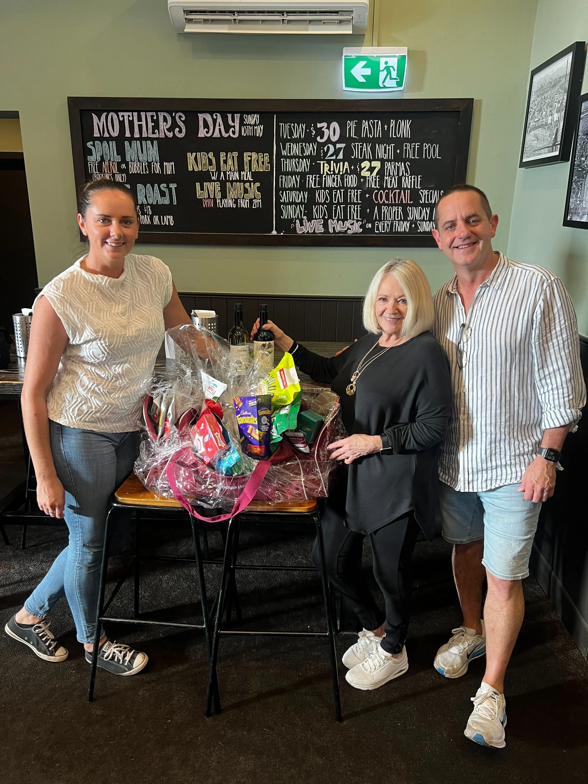 A group of three people, two women and one man, are standing around a table with a gift basket filled with snacks and wine, inside a restaurant. A blackboard behind them displays a Mother's Day menu and event details. The woman on the left is wearing