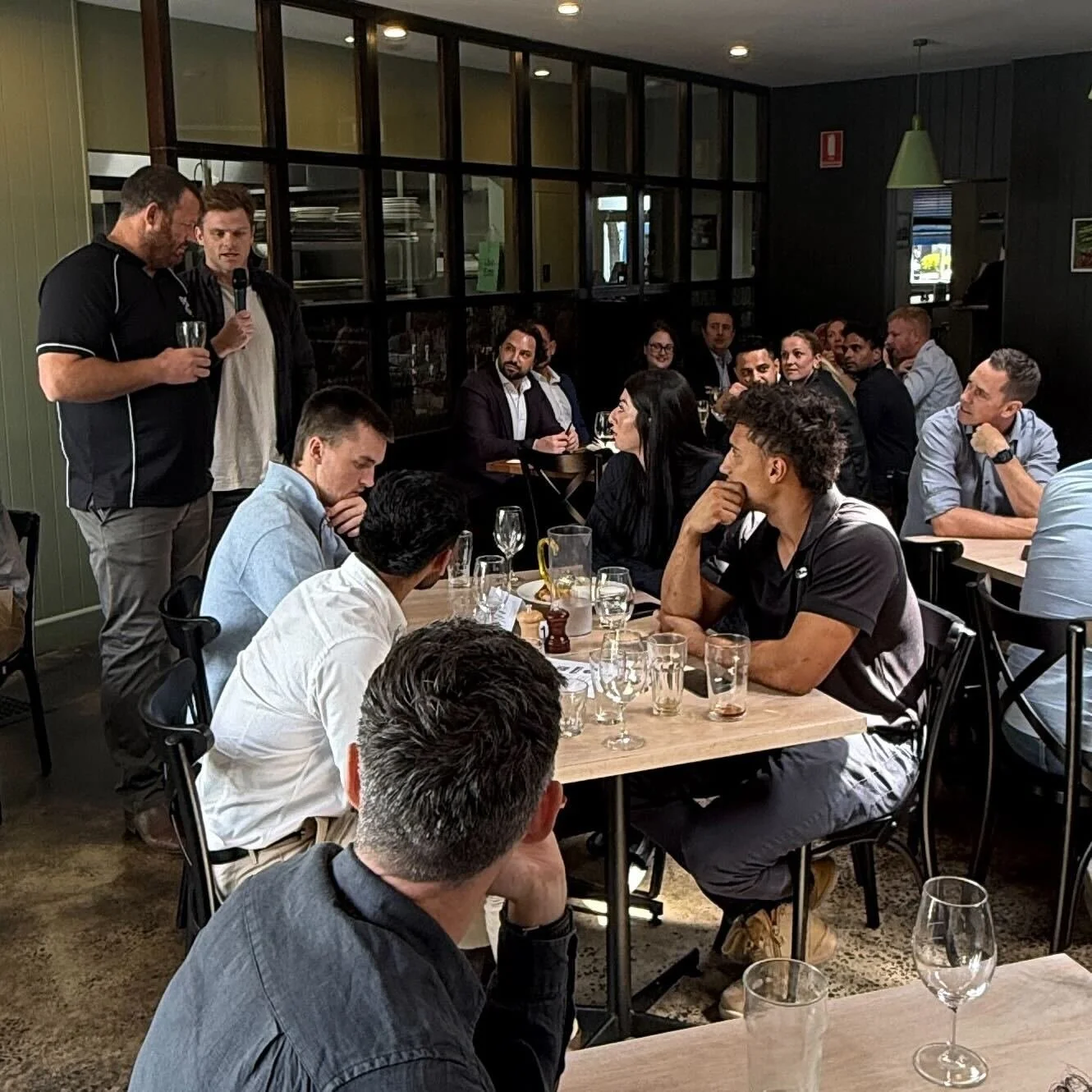 Group of people gathered in a restaurant for a presentation or speech, with two men standing at the front, one holding a glass and the other speaking into a microphone.