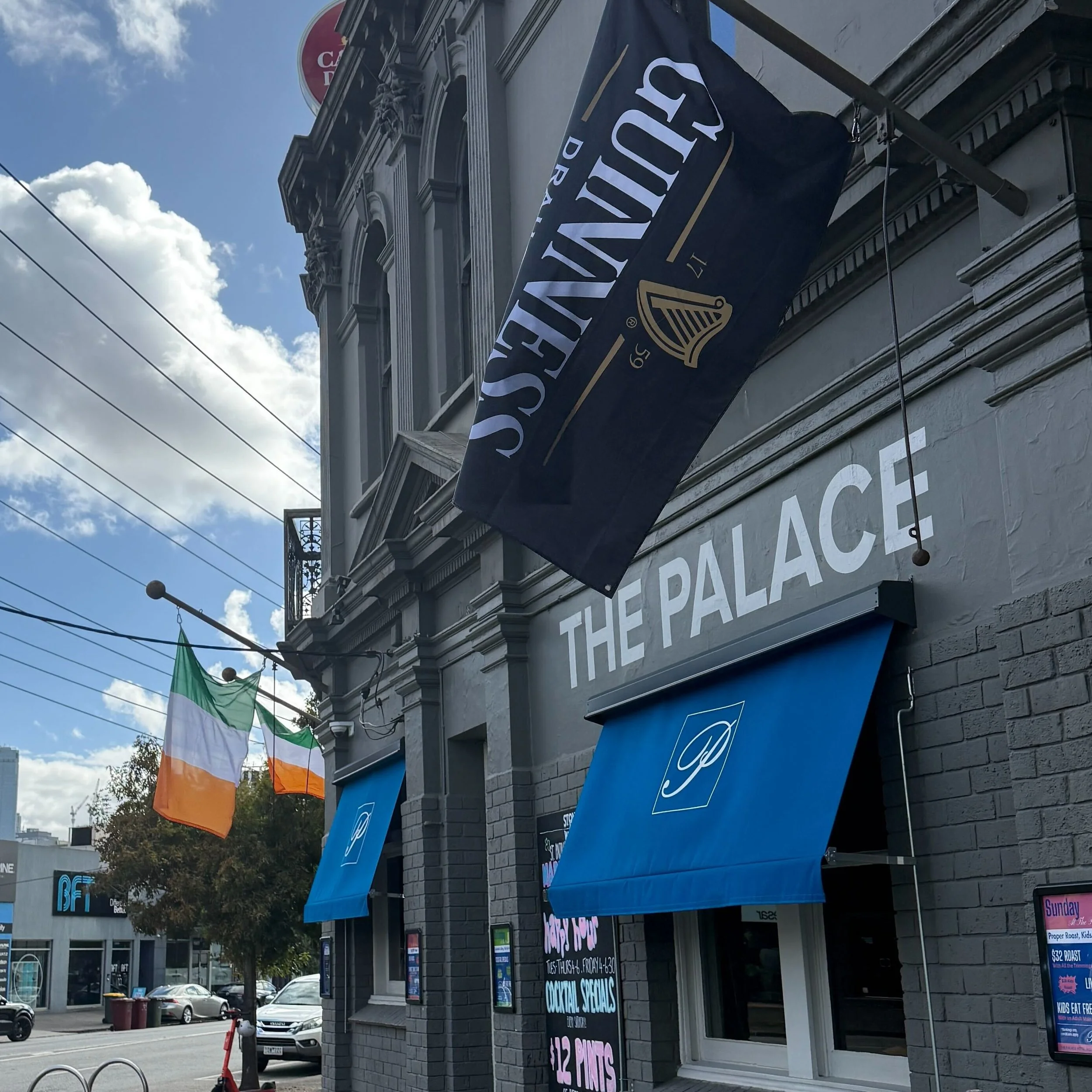 Street view of a building with ornate architectural details, displaying banners and signs for a bar and restaurant, with an Irish flag hanging nearby under a partly cloudy sky.