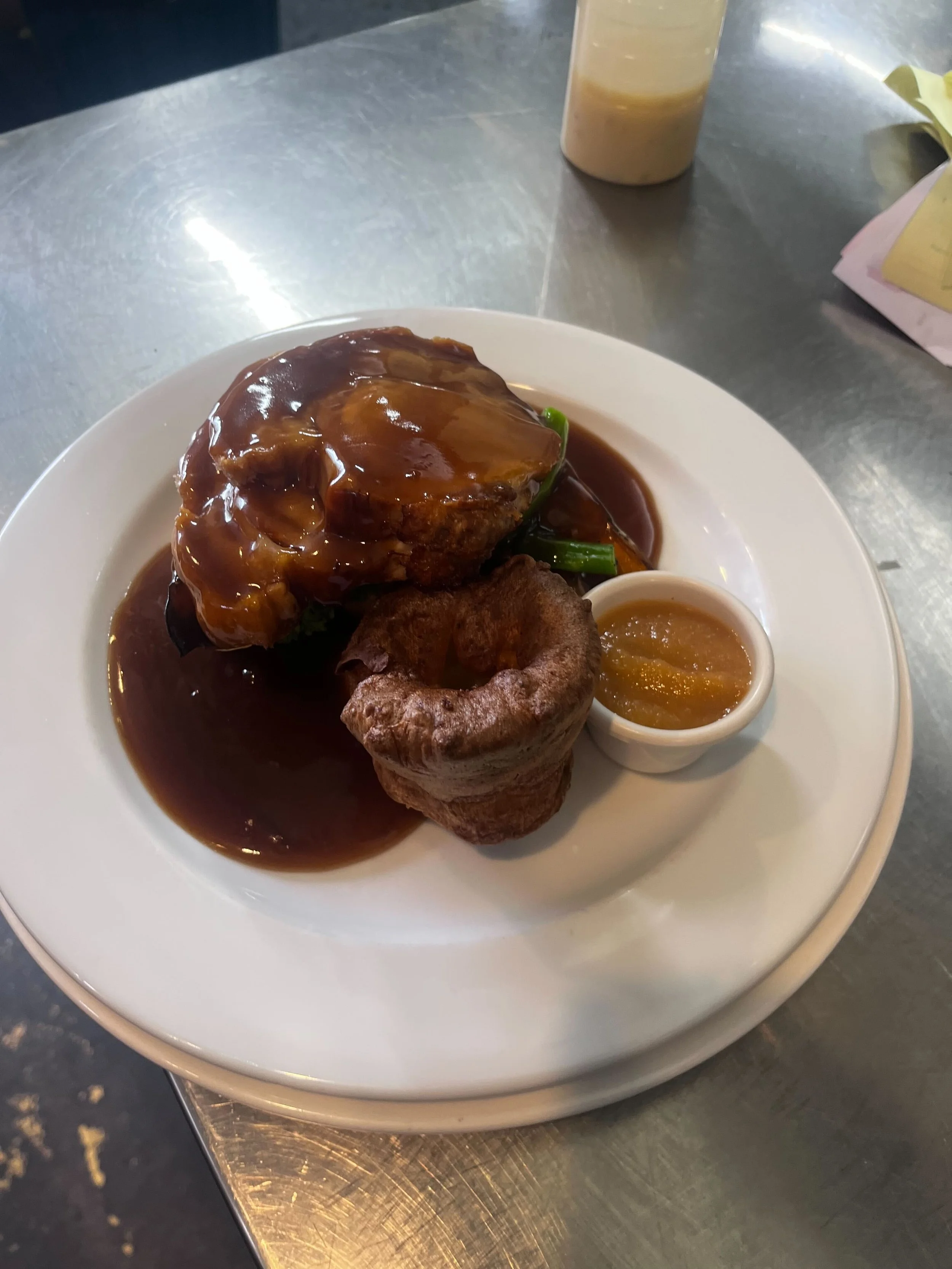 A plate with a large piece of meat topped with brown gravy, a small cup of yellow sauce, a fried roll, and some green vegetables. In the background, a drink container and a paper bag are visible on a metal table.