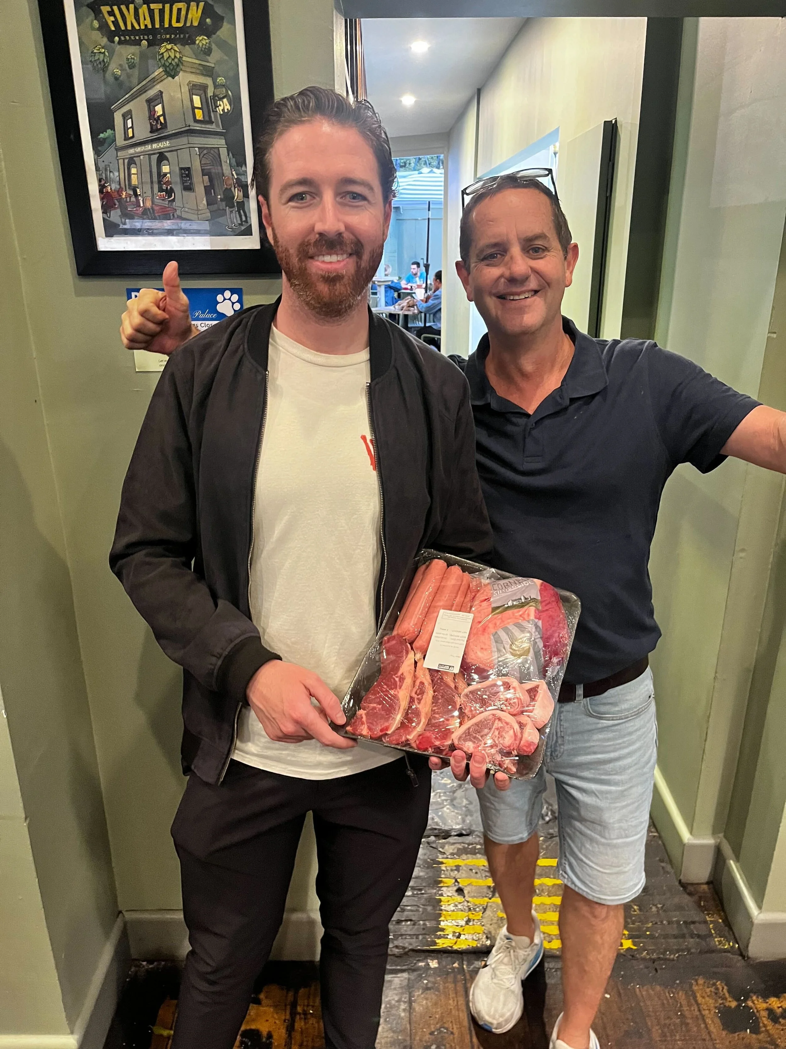 Two men smiling and standing in a hallway, one holding a tray of raw meat. The man on the left has a beard, is wearing a black jacket, and the man on the right is wearing a navy polo shirt and glasses on his head. There is a framed poster on the wall