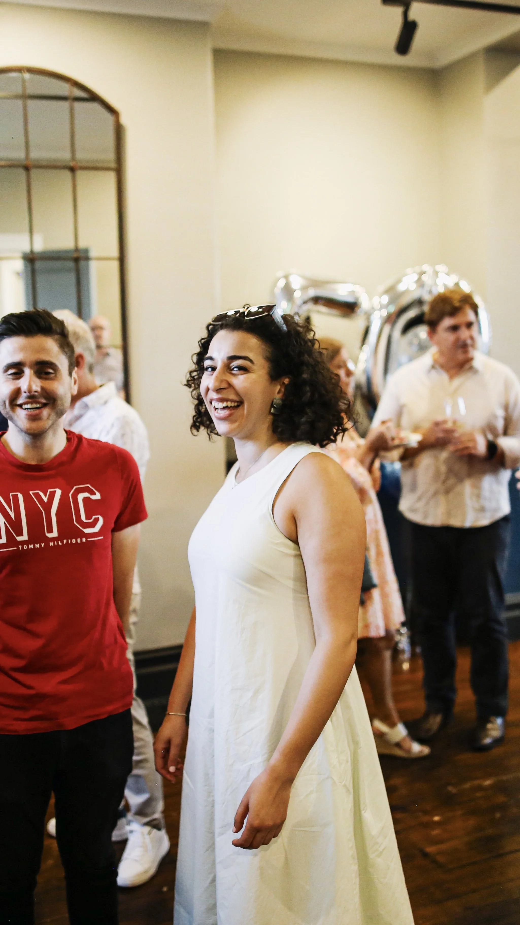 Smiling woman in a cream dress attending party upstairs at the Palace Hotel with people in the background, including a man in a red shirt and other guests with balloons.